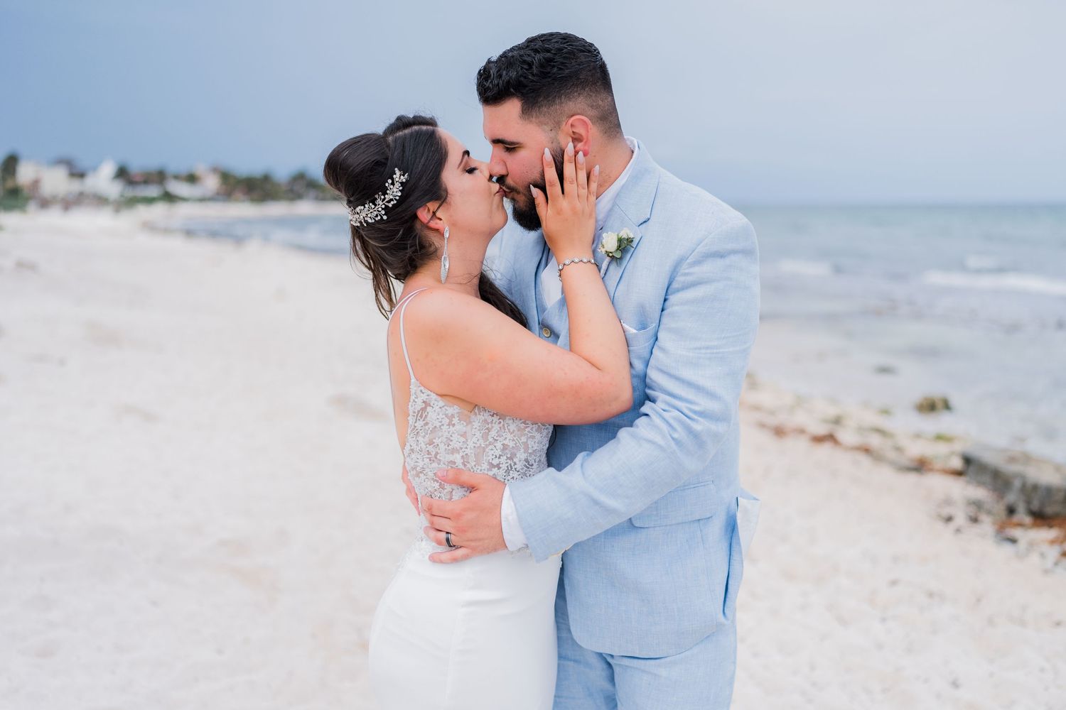 Bride and bearded groom share a kiss on a white sand beach, bride wearing floral hair accessory.