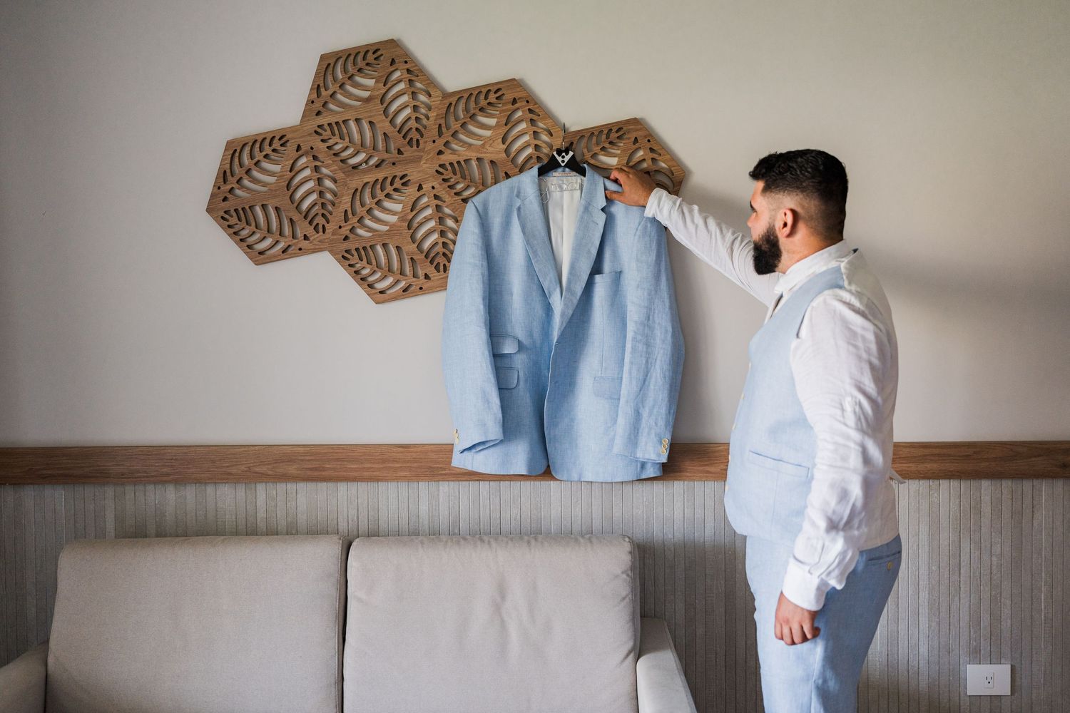 Dapper bearded groom holding light blue suit jacket on hanger in hotel room before wedding.