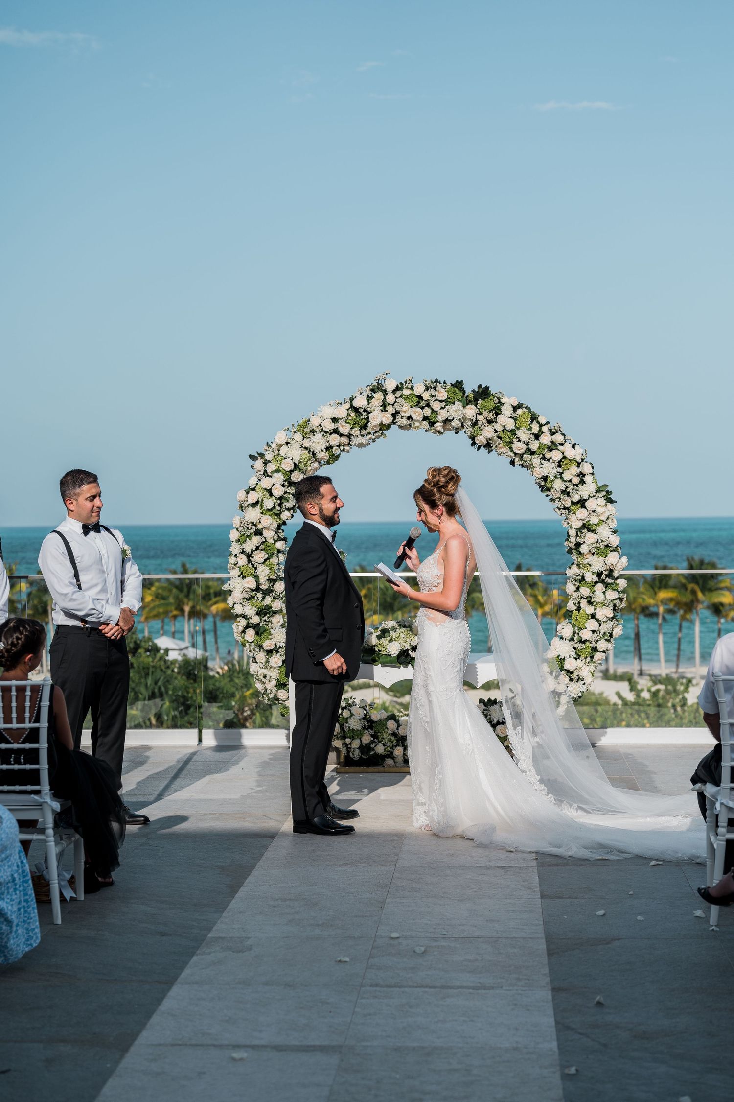 Bride and groom exchange vows under white floral circular arch with ocean backdrop.