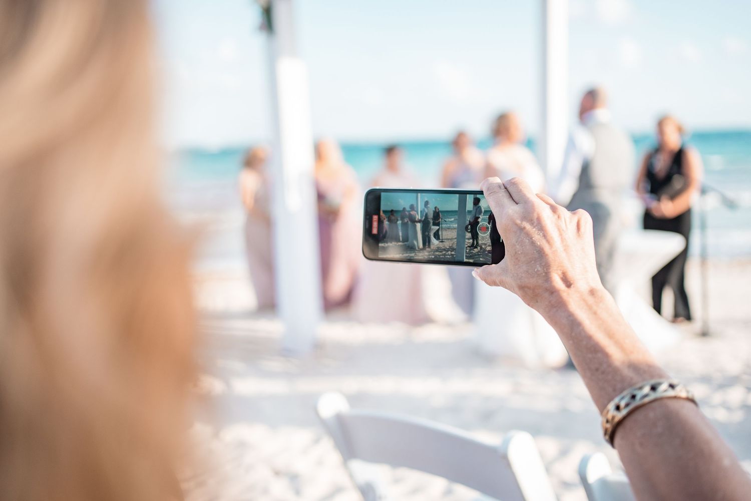 Guest captures beach wedding ceremony on smartphone with bridal party and ocean visible in background.
