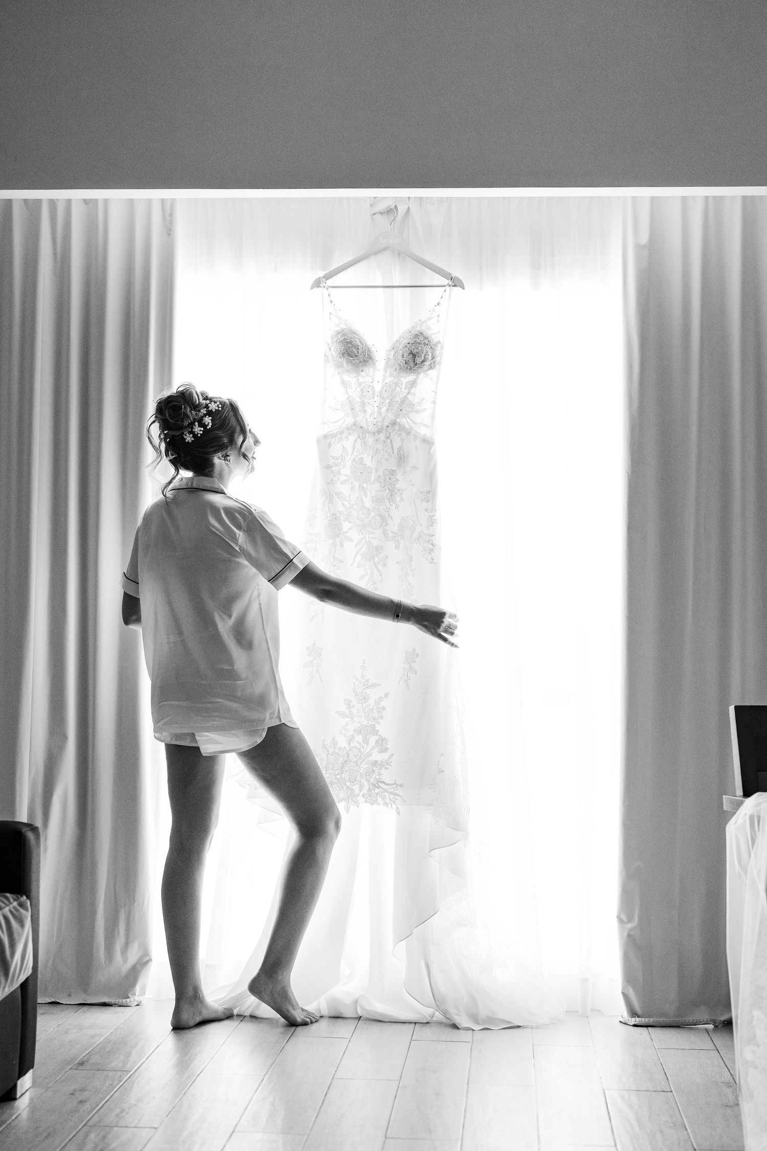 Black and white photo of bride reaching for wedding dress hanging in window.