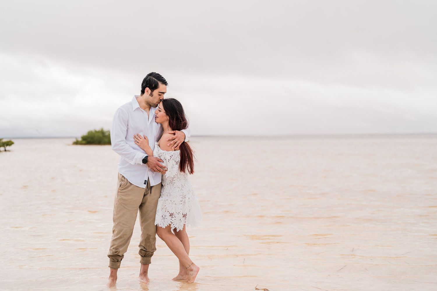 Romantic couple embracing barefoot in shallow ocean water on a cloudy beach during an engagement photoshoot.