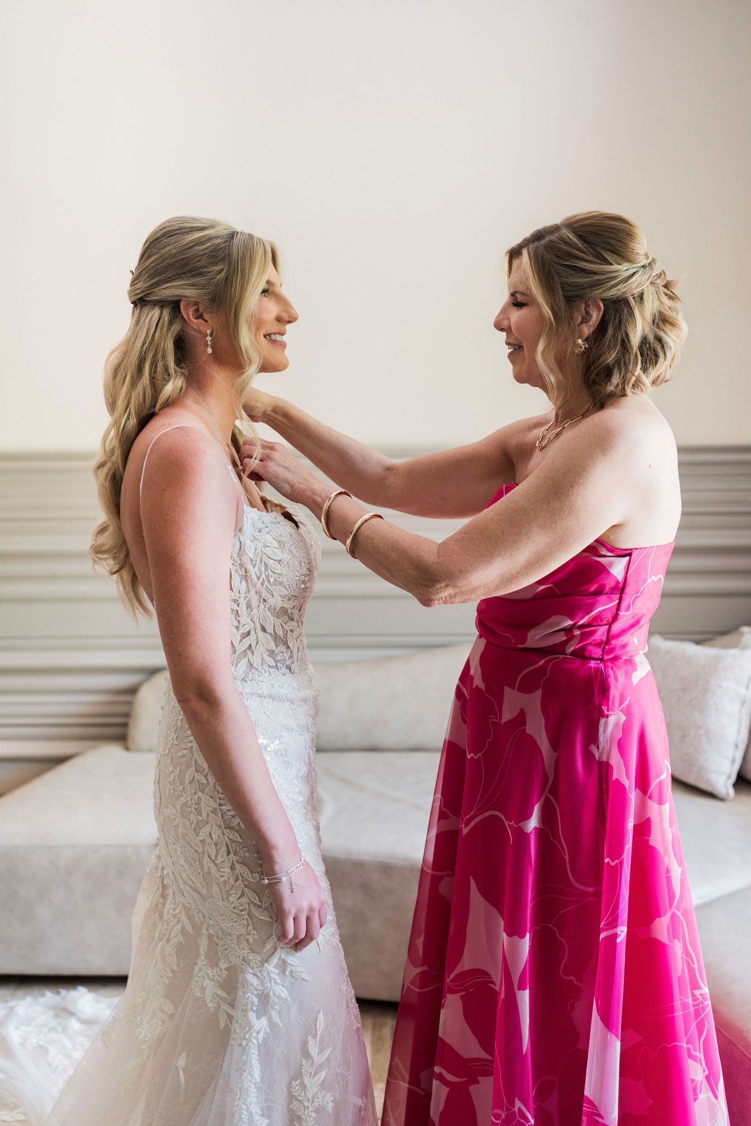 A bride in a lace gown smiles as a woman in a pink floral dress fastens her necklace in a bright room.