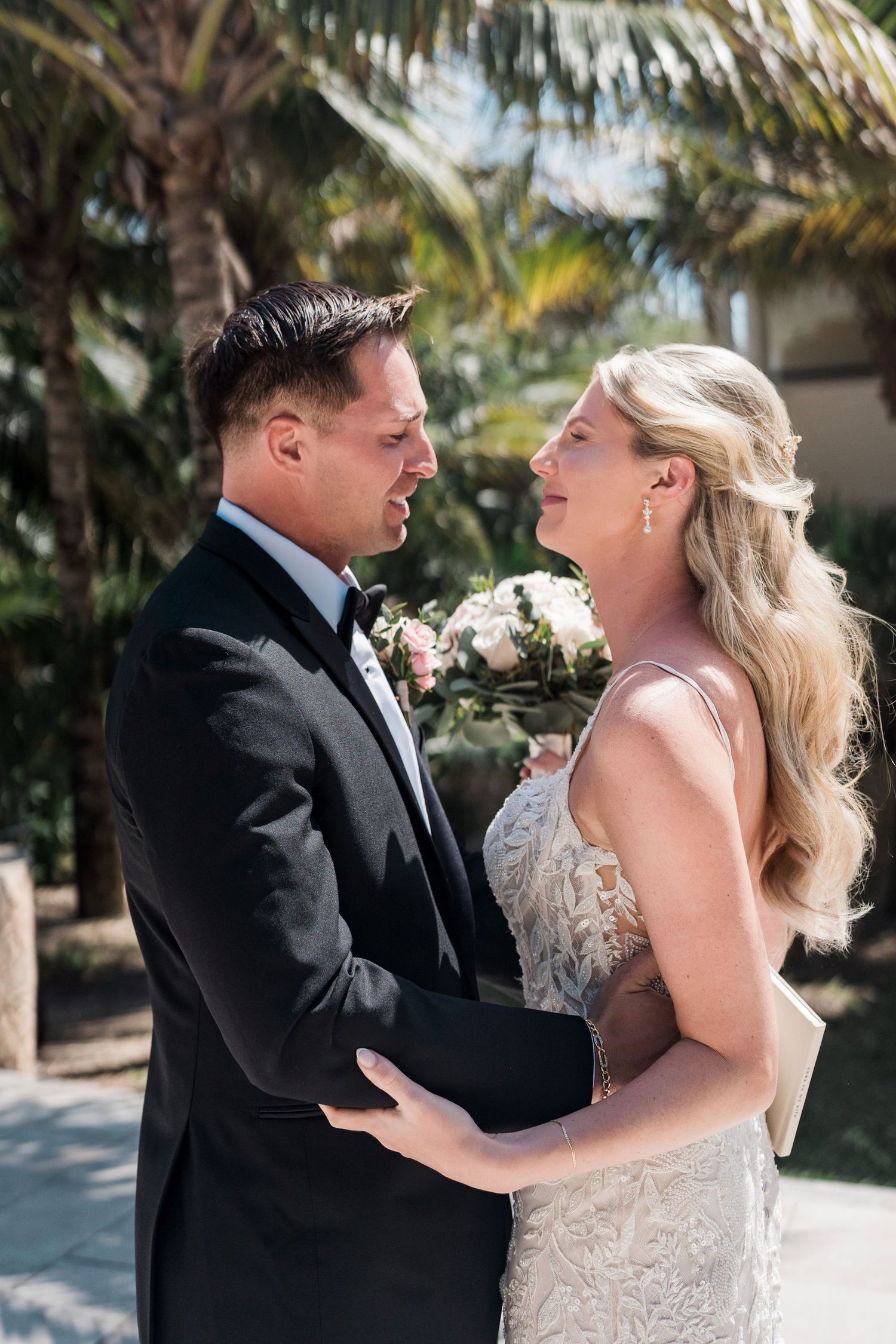 A bride and groom share a joyful moment outdoors, with the bride holding a bouquet and palm trees in the background.