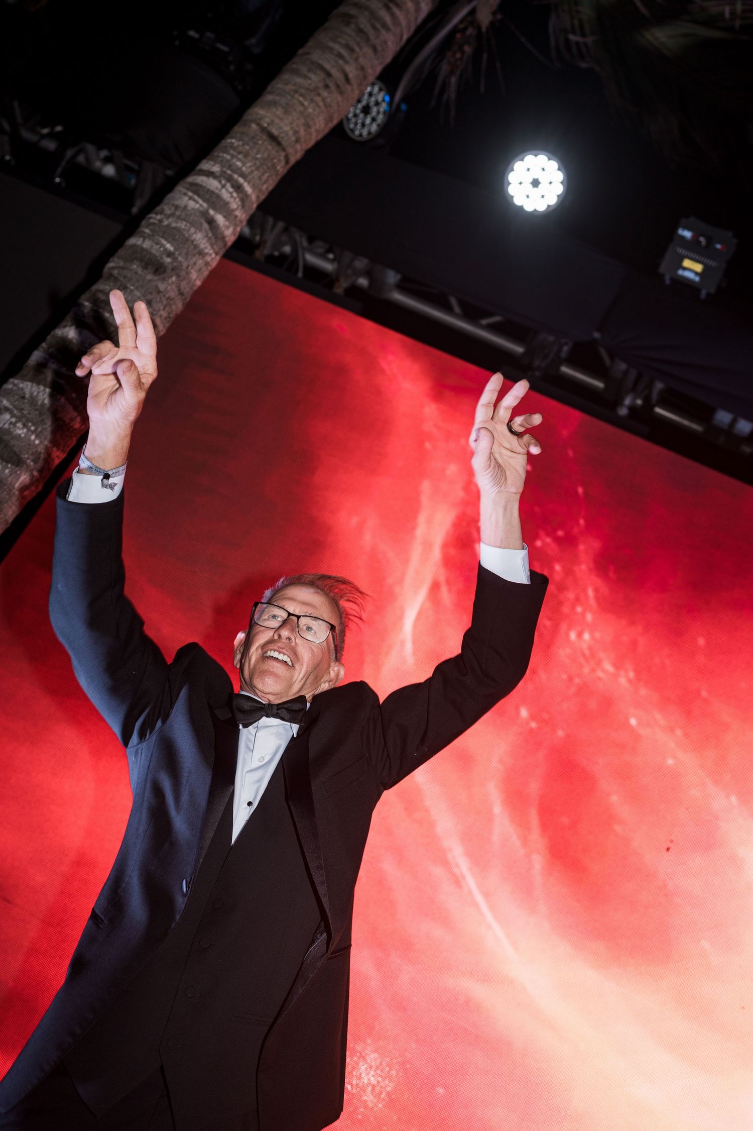 Man in tuxedo celebrating on stage with arms raised, red LED backdrop and stage lighting at formal event.