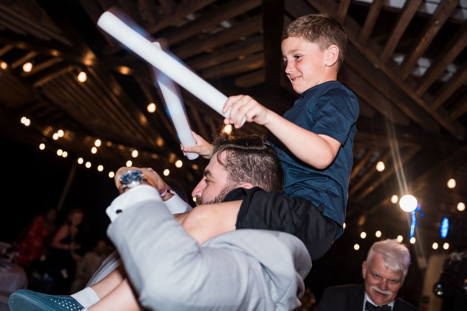 Boy holding glow stick sits on man's shoulders at festive indoor wedding reception with string lights.