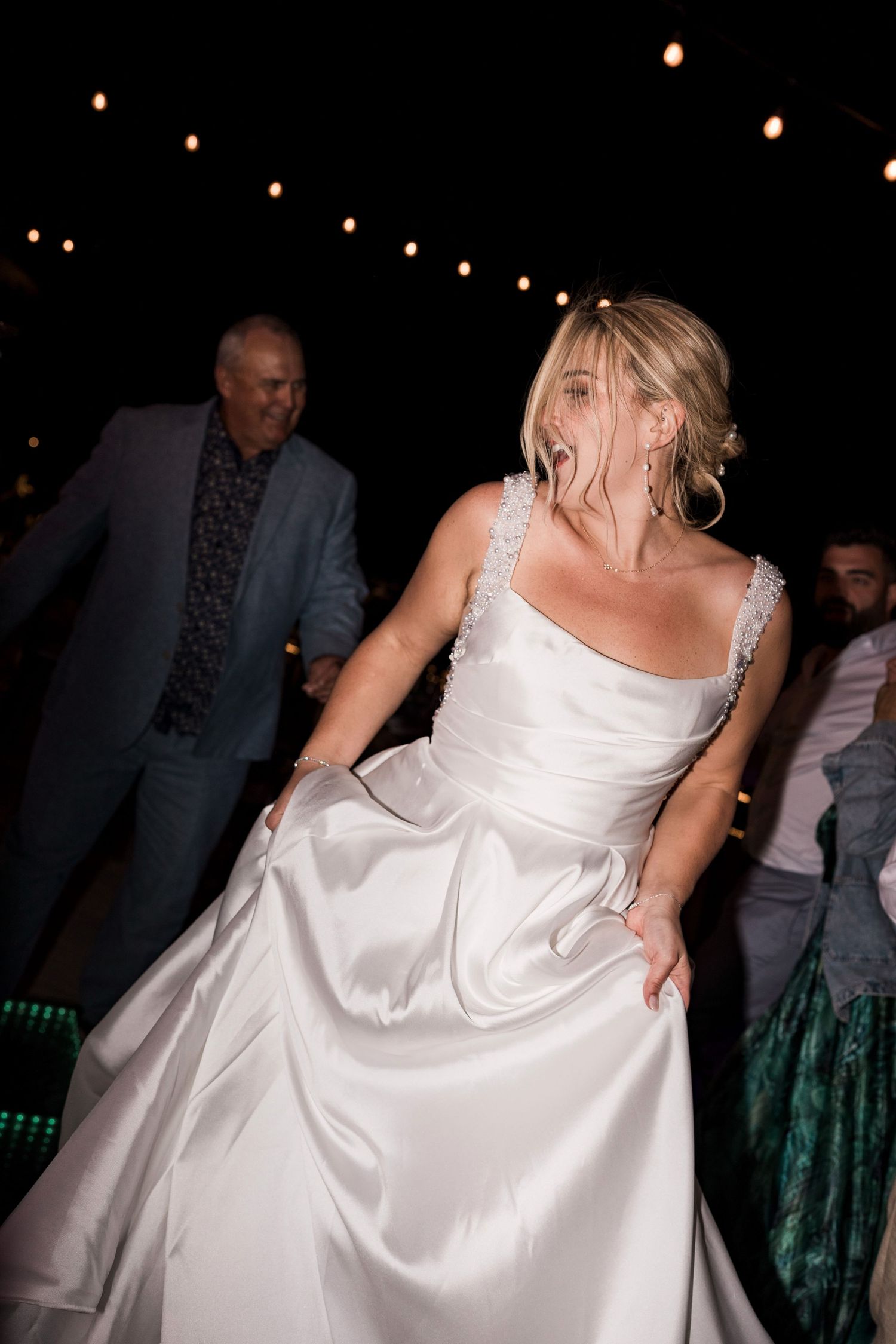 Bride in white satin gown dancing joyfully at outdoor wedding reception under string lights at night.