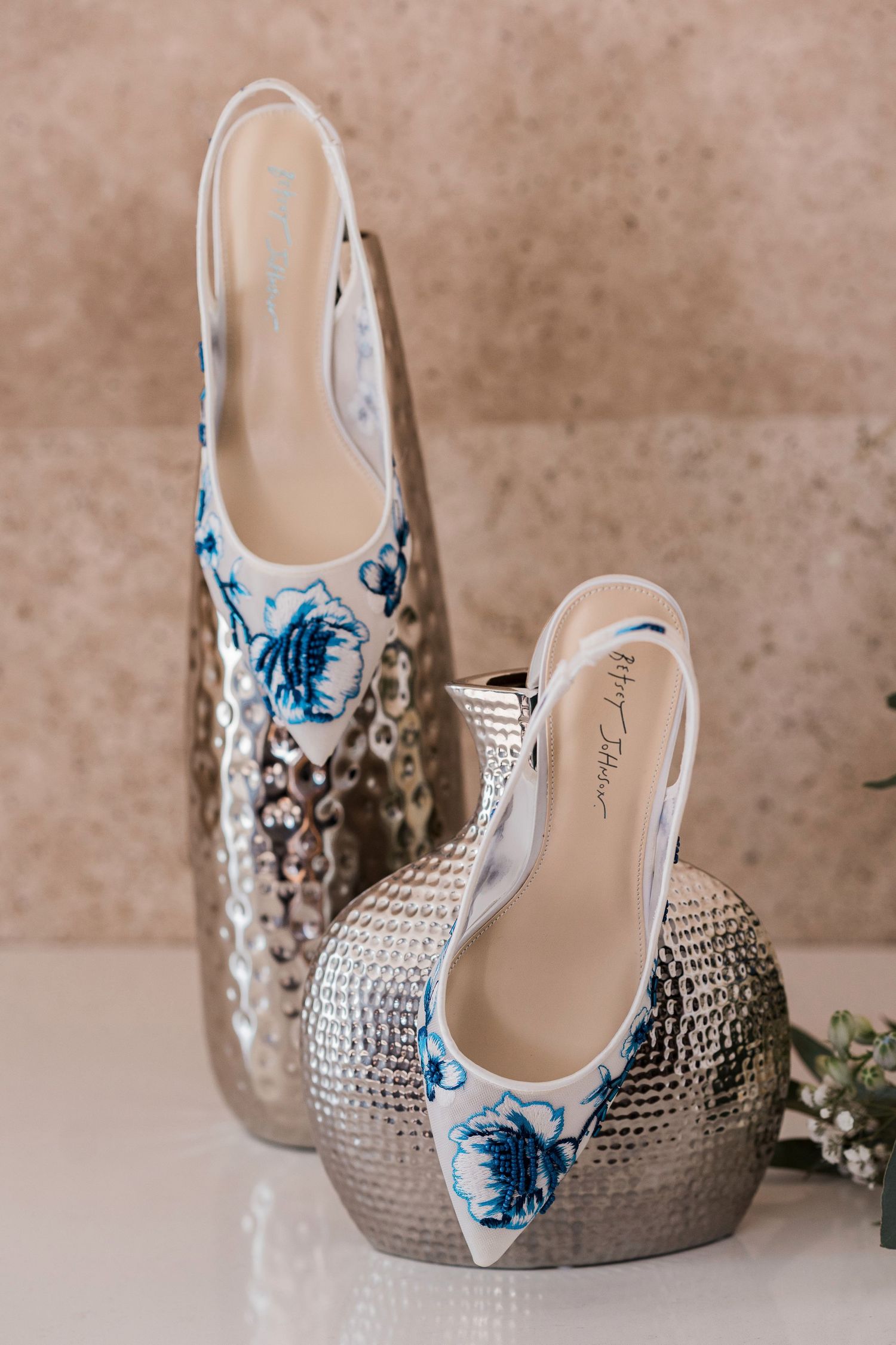 White slingback heels with blue floral embroidery displayed on silver metallic vases against a beige marble background.