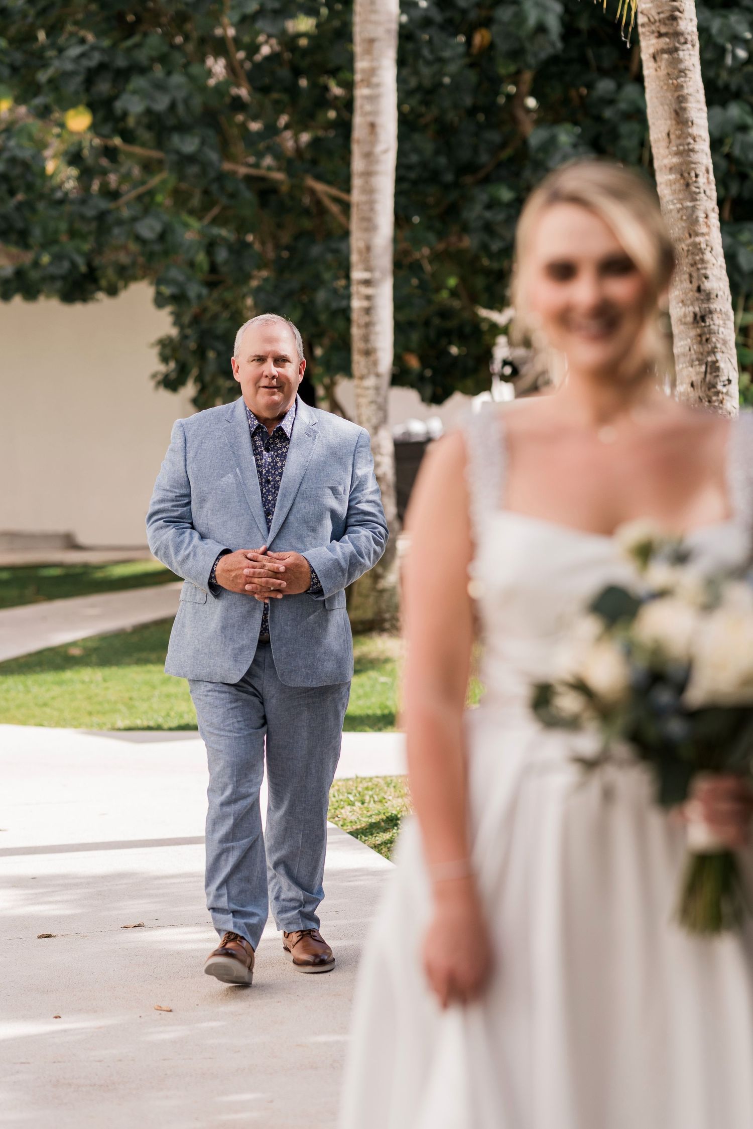 A bride in a white gown holds a bouquet as her father walks toward her during an outdoor wedding first look.