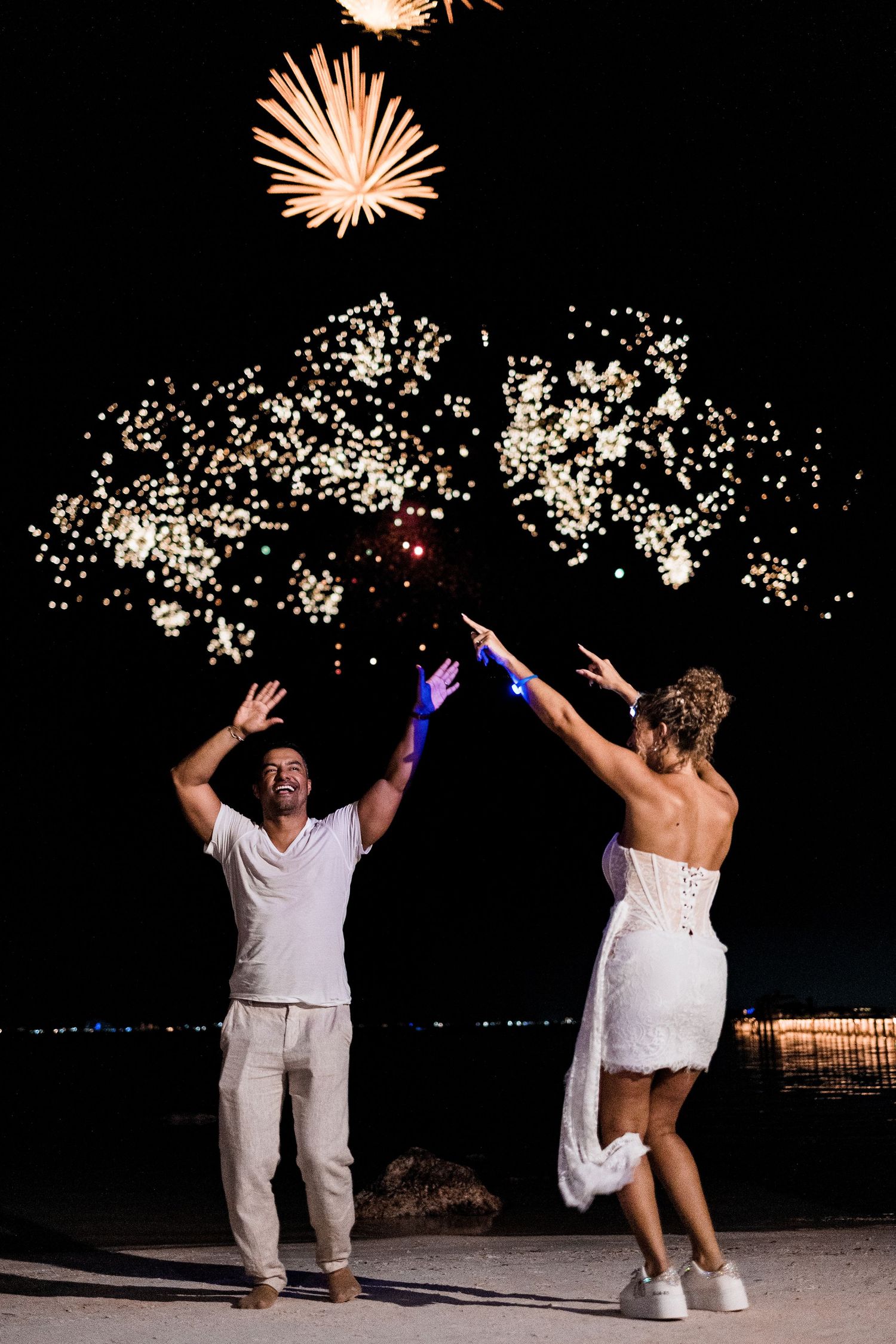 Couple in white celebrating with fireworks lighting up the night sky at a beach wedding reception. Puerto Vallarta wedding photographer
