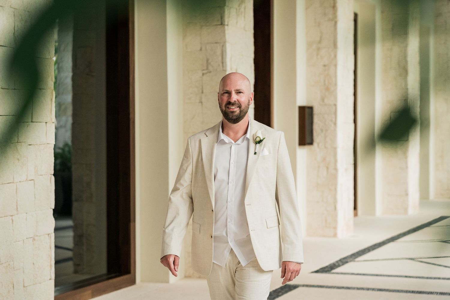 Smiling groom in a light beige suit with a floral boutonniere stands in a sunlit outdoor corridor.