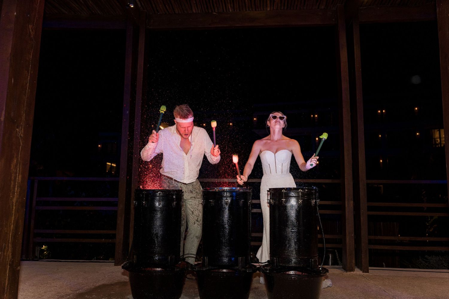 Bride and groom playing large drums together at an outdoor wedding reception at night.