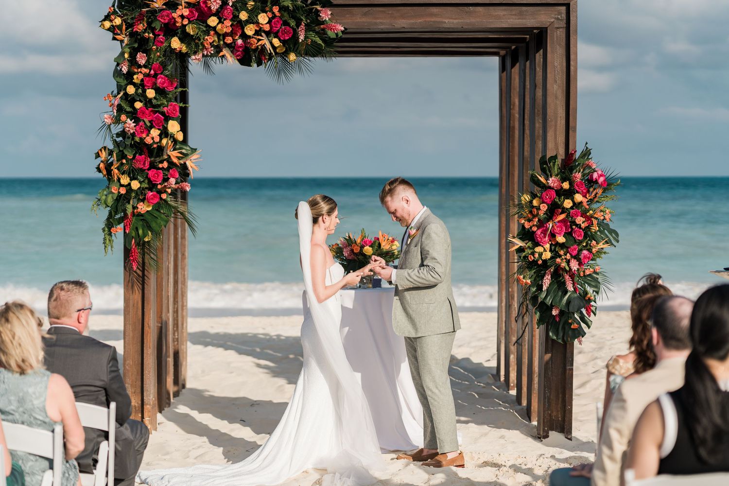 Couple exchanging vows under a floral wooden arch on a tropical beach wedding ceremony with colorful blooms.
