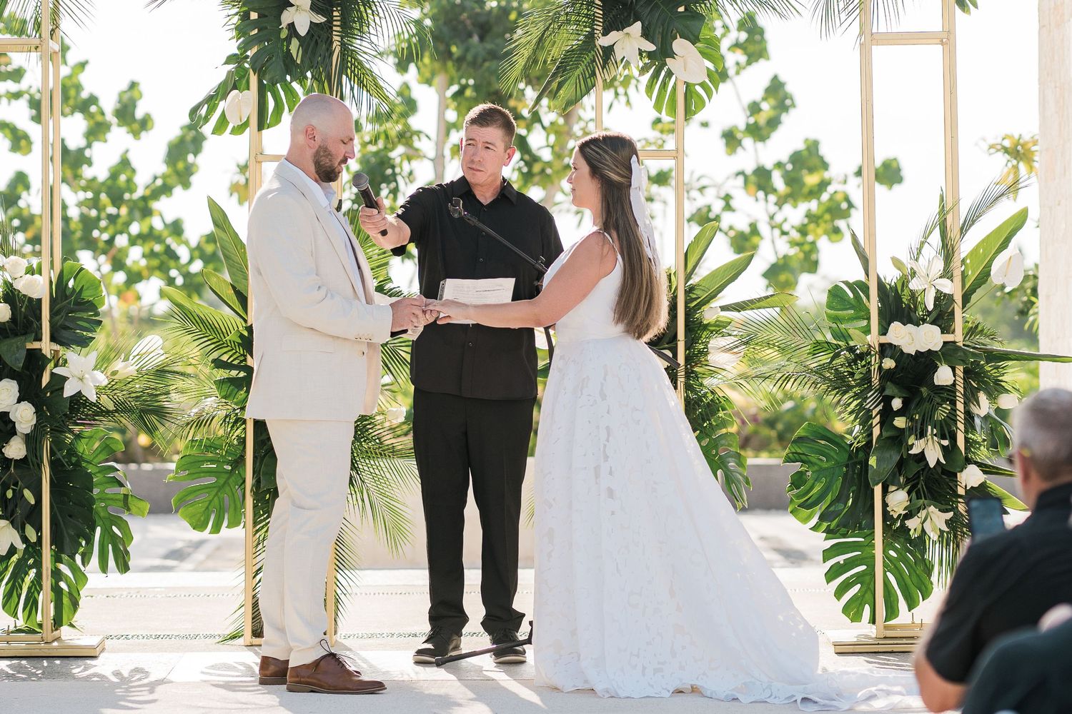 Couple exchanging vows at outdoor wedding ceremony with tropical greenery and white floral arch backdrop.
