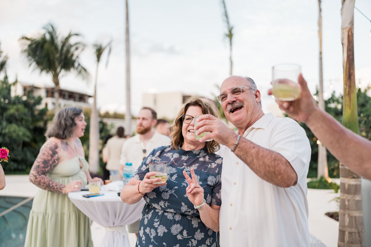 Couple laughing and holding drinks at an outdoor tropical wedding reception party near a pool.