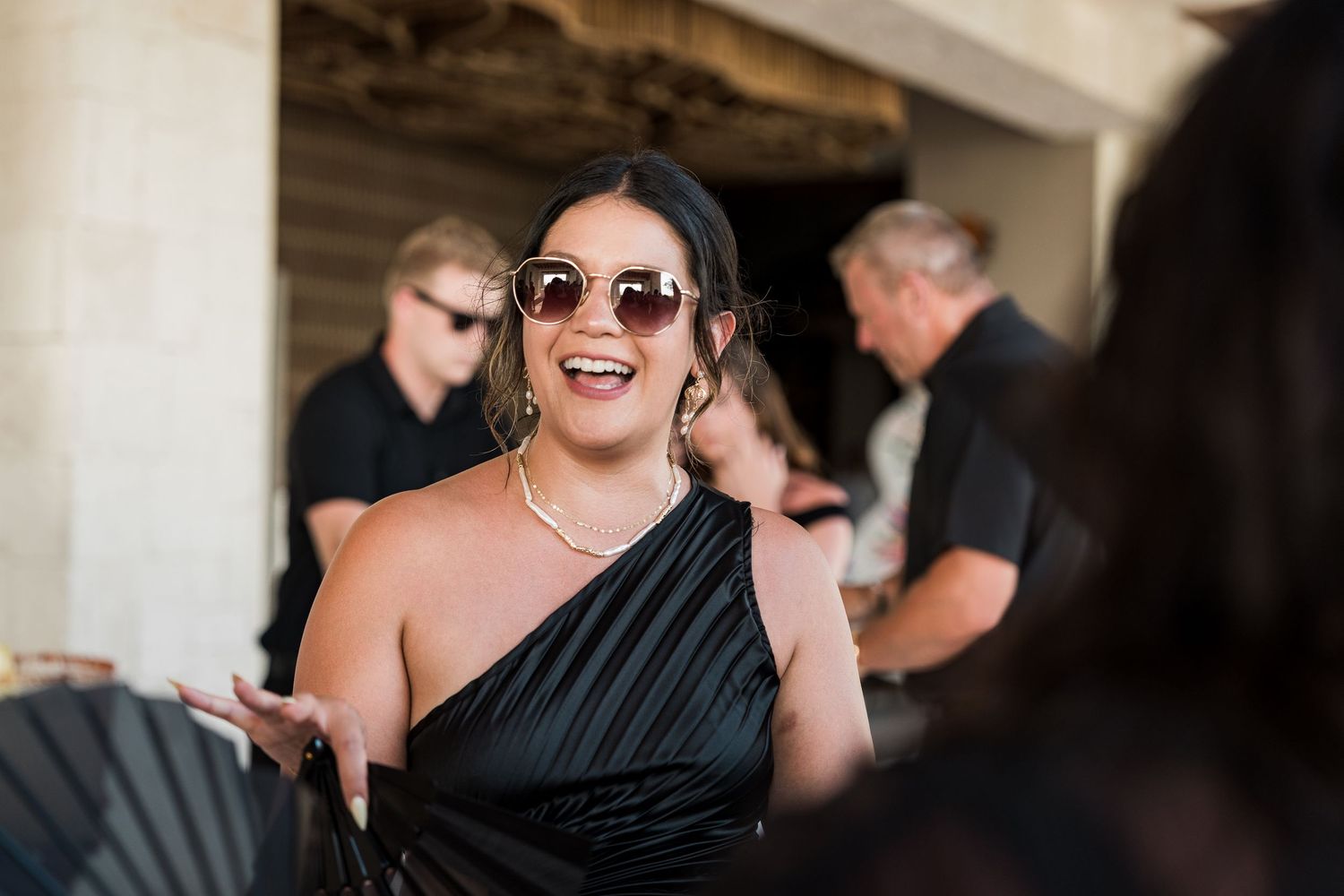 Woman in black one-shoulder dress and sunglasses smiling at an outdoor event.