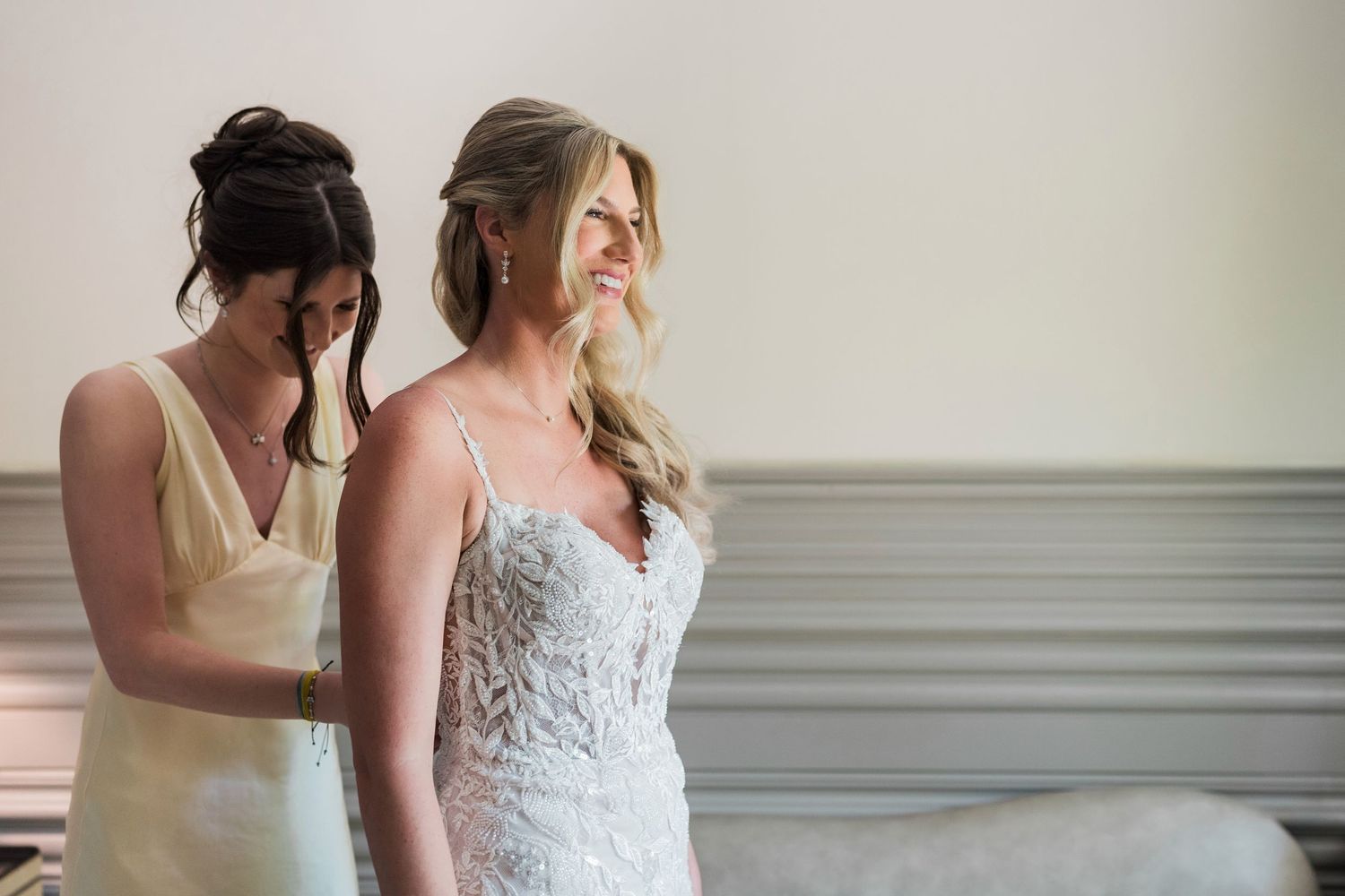 Bride in lace wedding gown being helped by bridesmaid in yellow dress during wedding preparation.