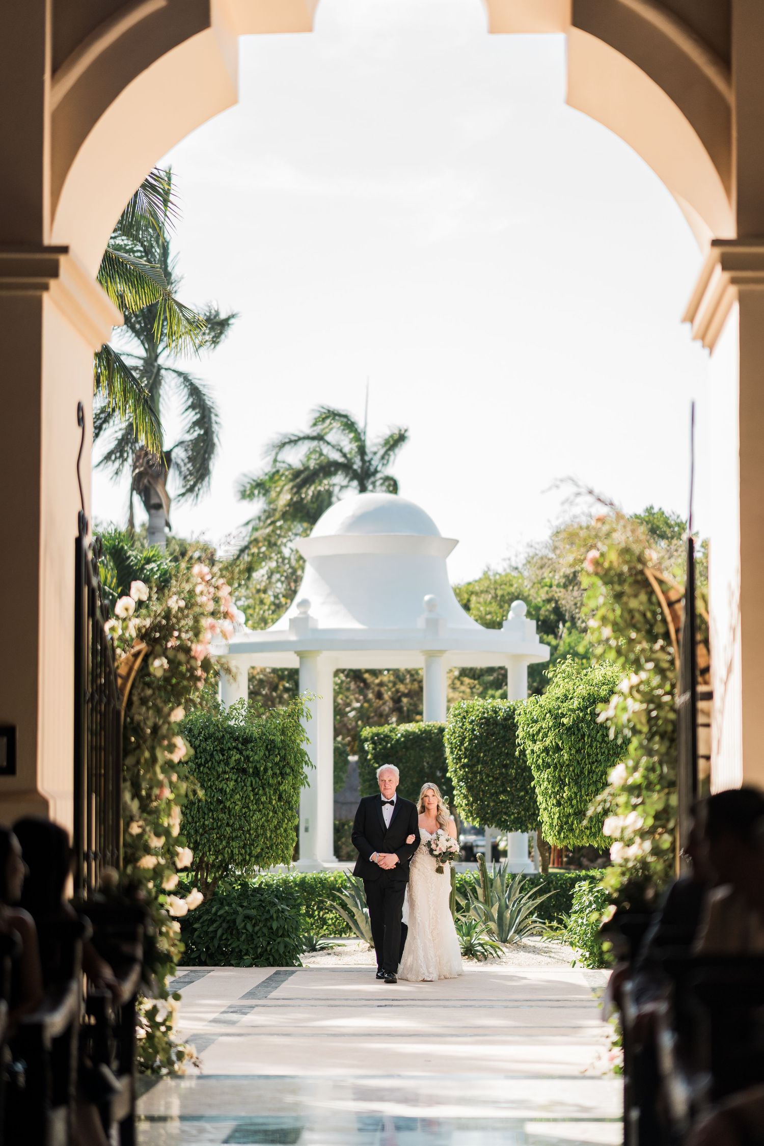 Bride and groom walk through elegant archway toward white gazebo surrounded by lush greenery at outdoor wedding ceremony.