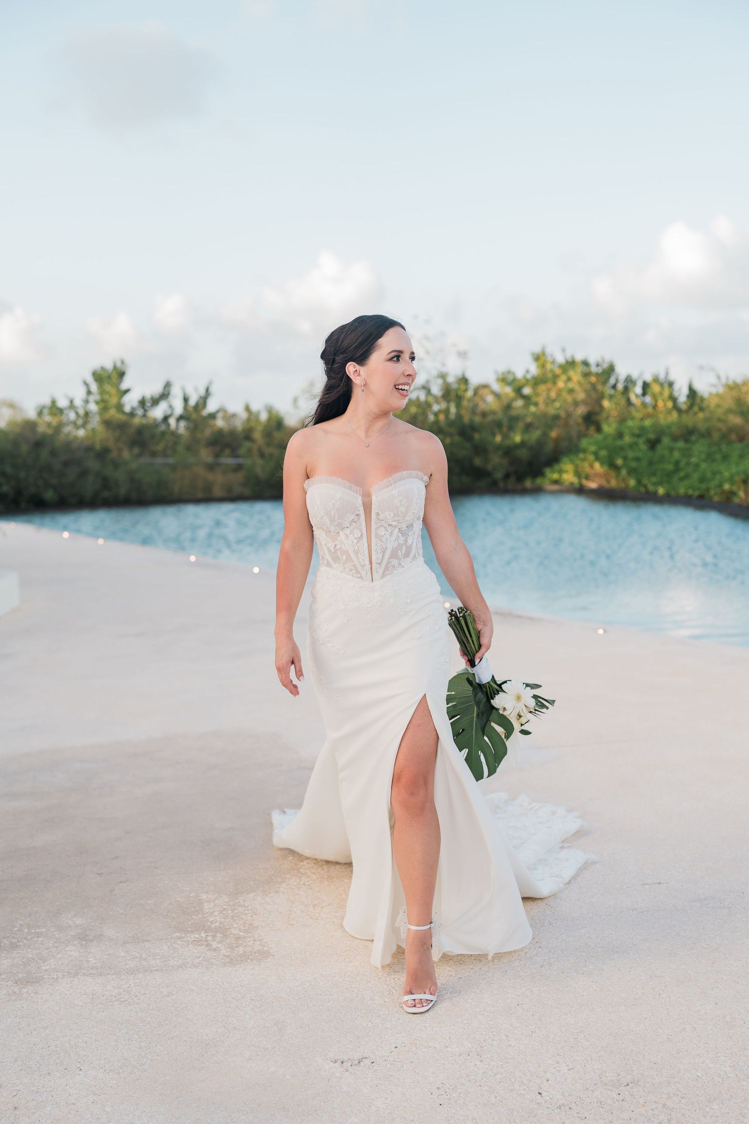 Bride in strapless lace wedding gown with slit holds tropical bouquet poolside at outdoor beach venue.