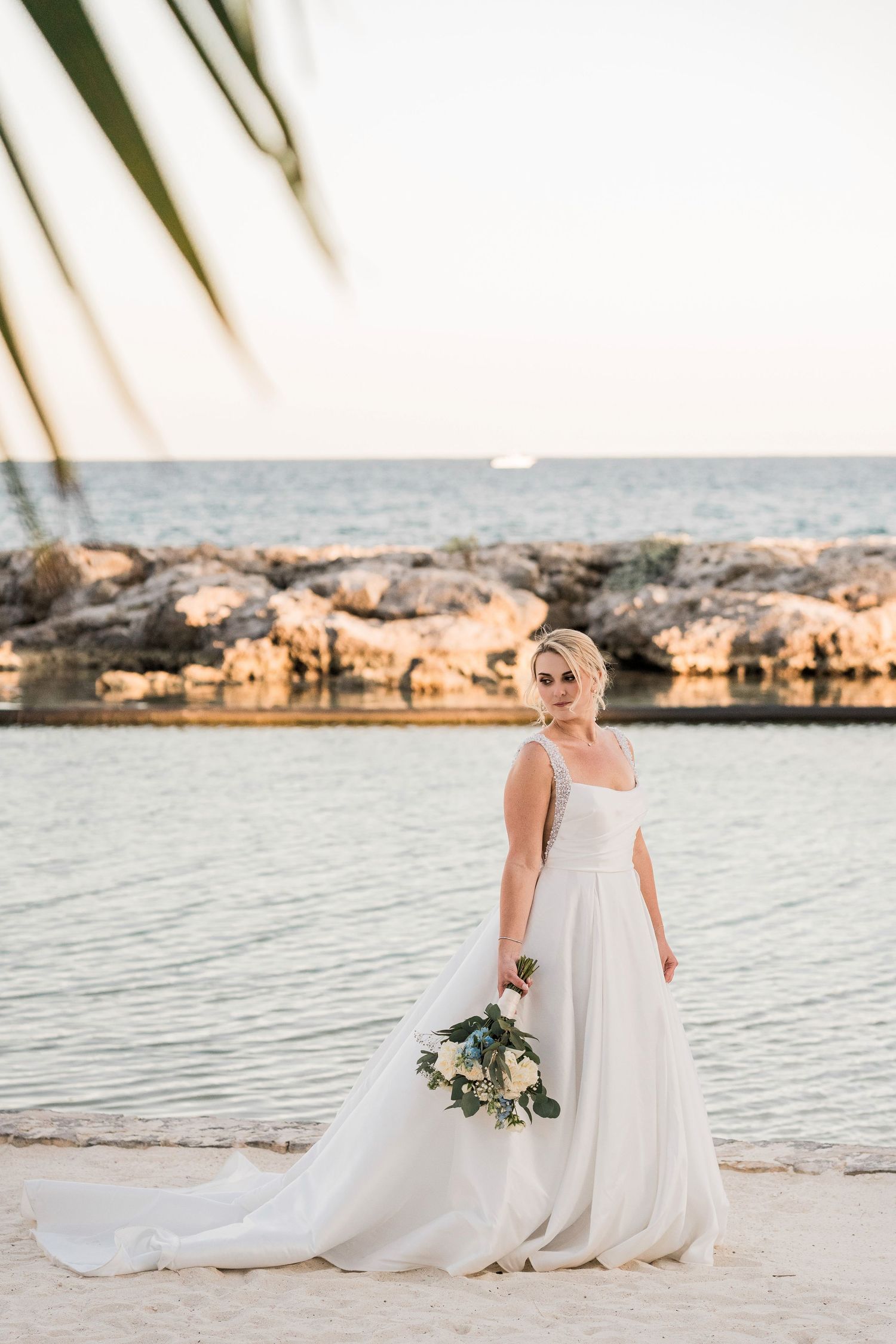 Bride in elegant white gown holding bouquet on sandy beach at sunset with rocky shoreline backdrop.
