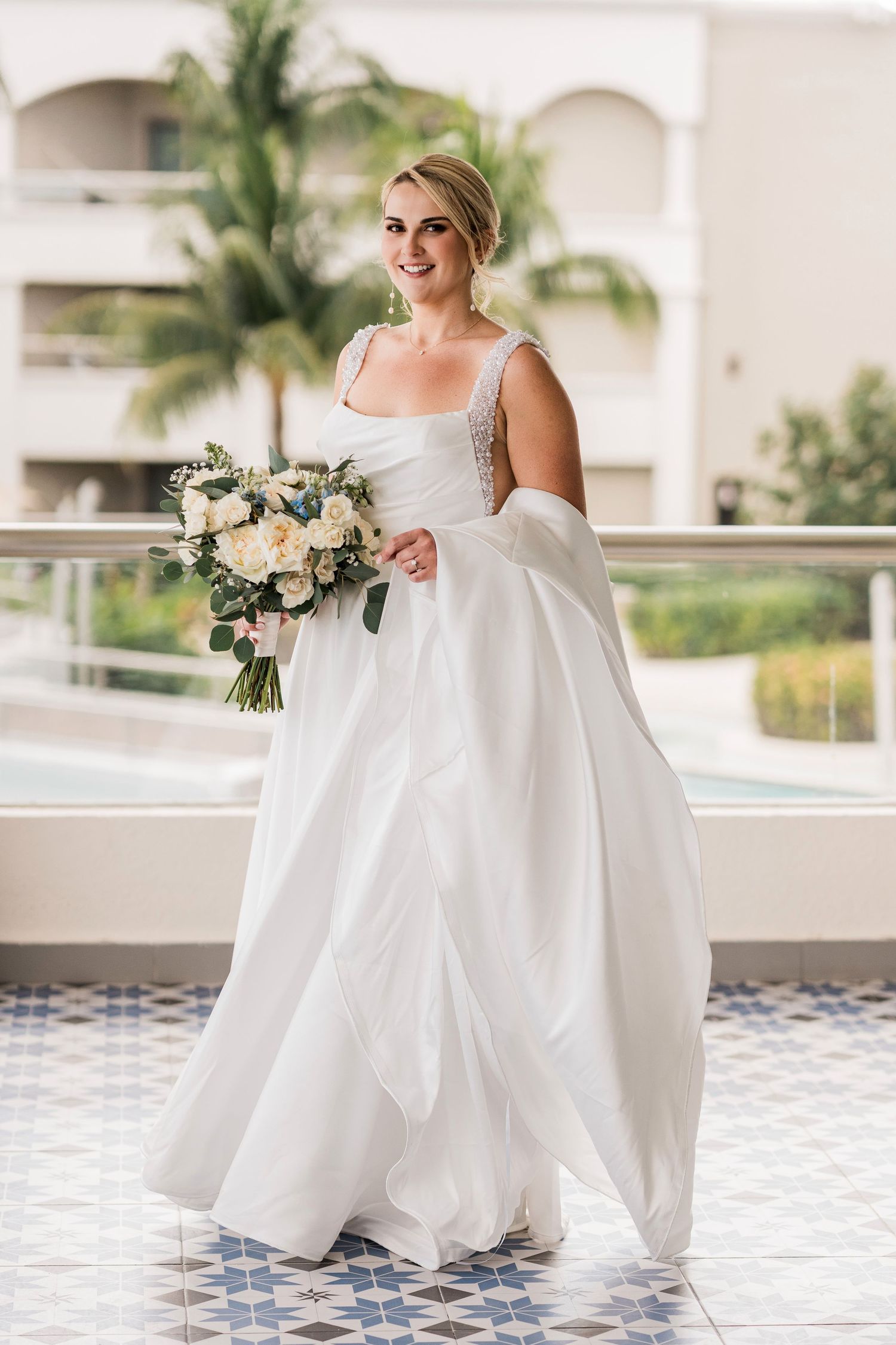 Smiling bride in a white ball gown holding a white and green bouquet on a balcony with palm trees.