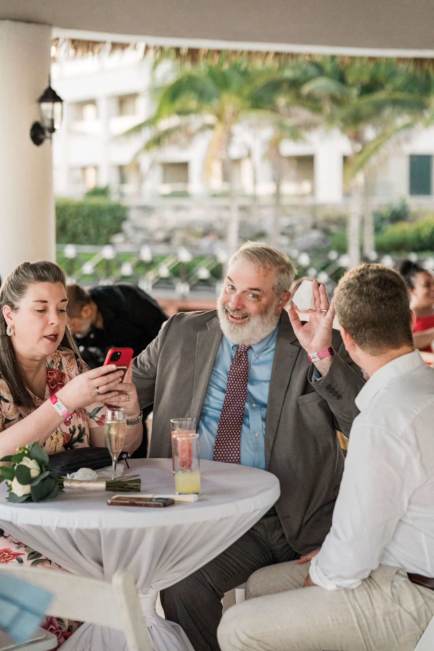 Guests laughing and sharing phones at an outdoor resort wedding cocktail hour with tropical palm trees.