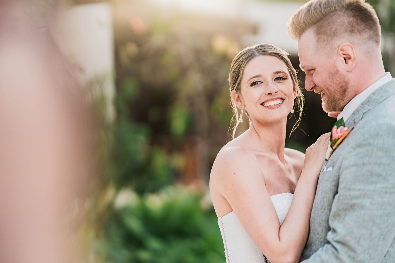 Bride smiling at groom in gray suit during outdoor wedding ceremony, surrounded by lush greenery in warm sunlight.