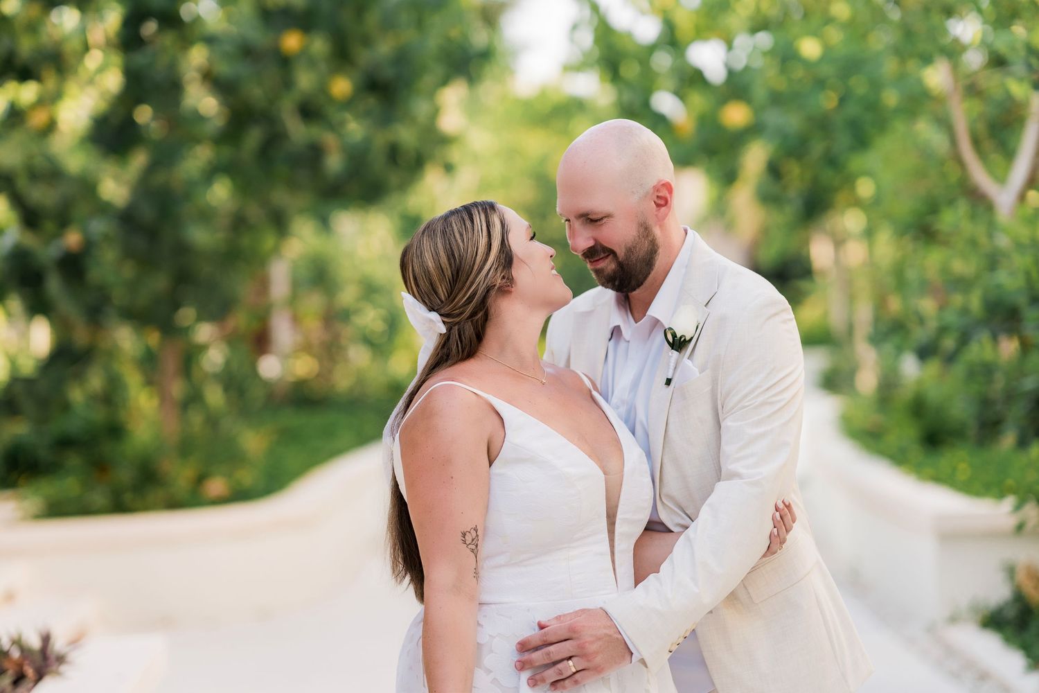 A bride and groom share a romantic moment outdoors, surrounded by lush greenery, as she wears a white gown and he a tan suit.