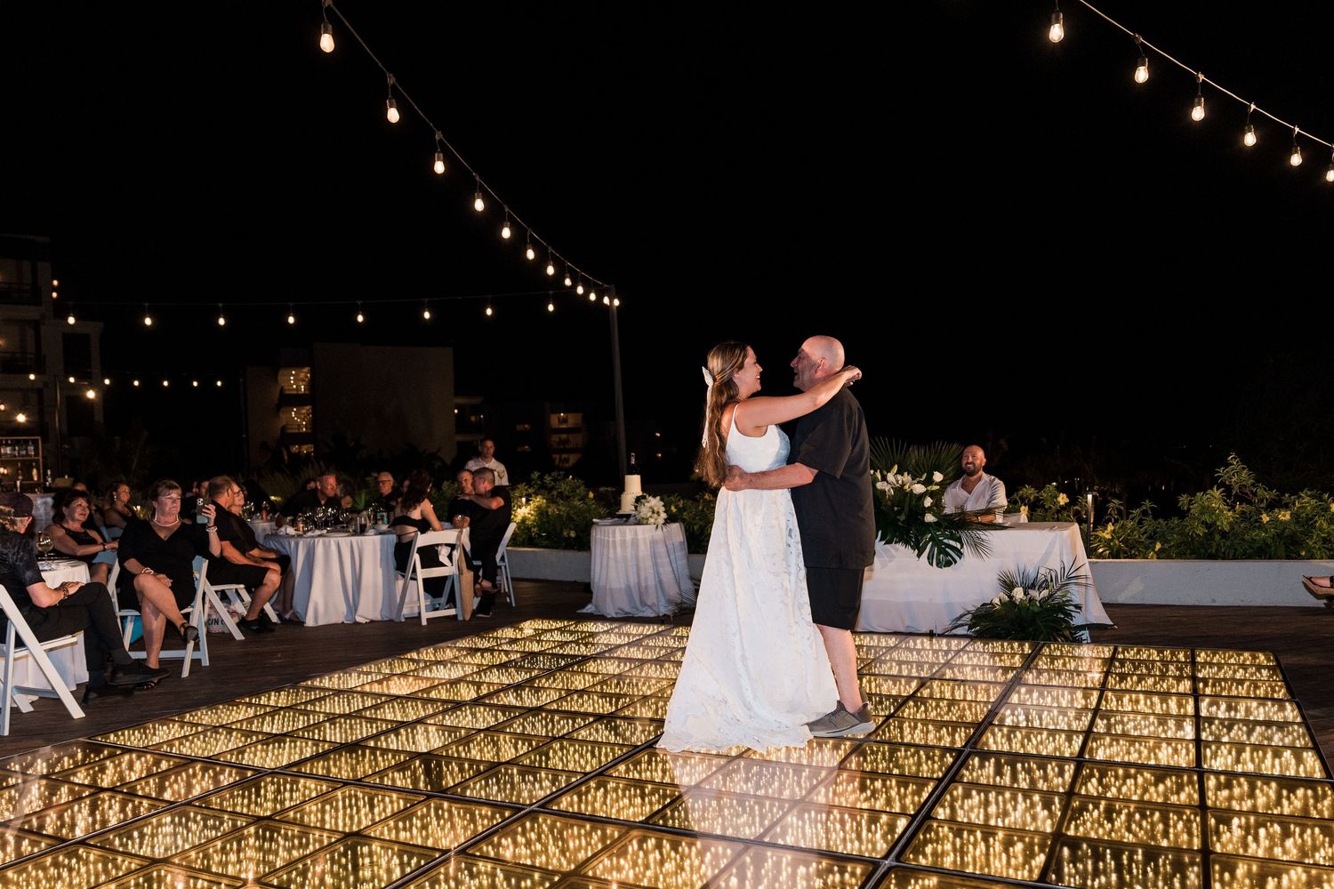 Bride and groom share first dance on illuminated floor under string lights at outdoor evening wedding reception.