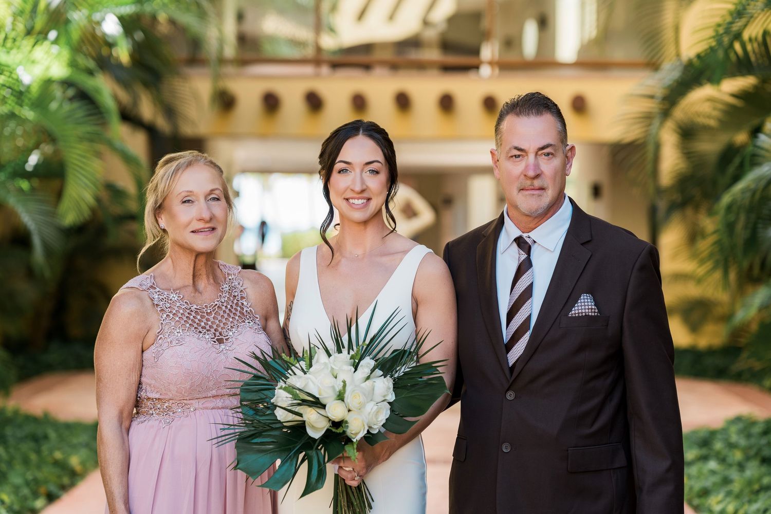 Bride in white gown holds tropical bouquet flanked by two guests in formal attire at an elegant venue.