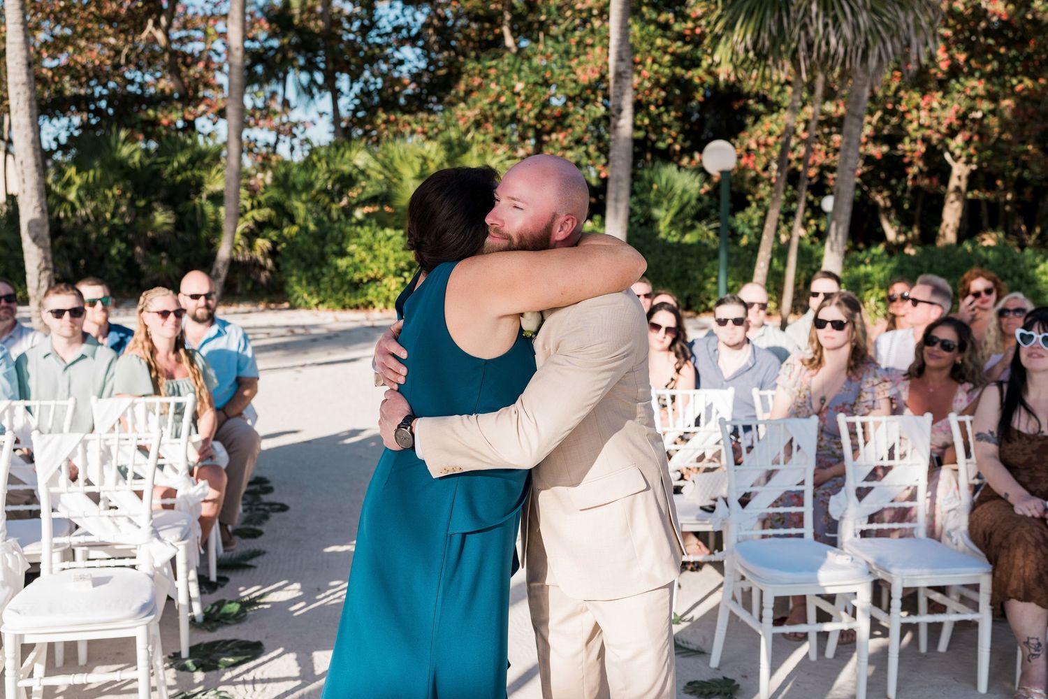 Bald groom in tan suit hugs woman in teal dress at outdoor beach wedding ceremony with white chairs.