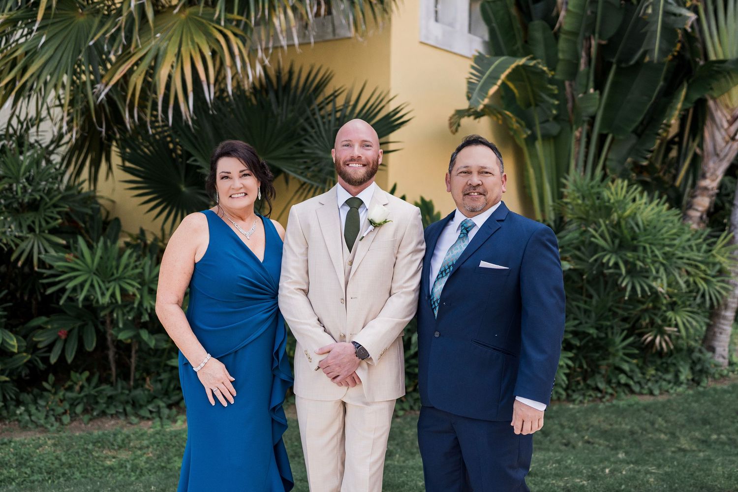 A bride in blue, groom in tan suit, and guest in navy pose outdoors near tropical palms at a wedding.