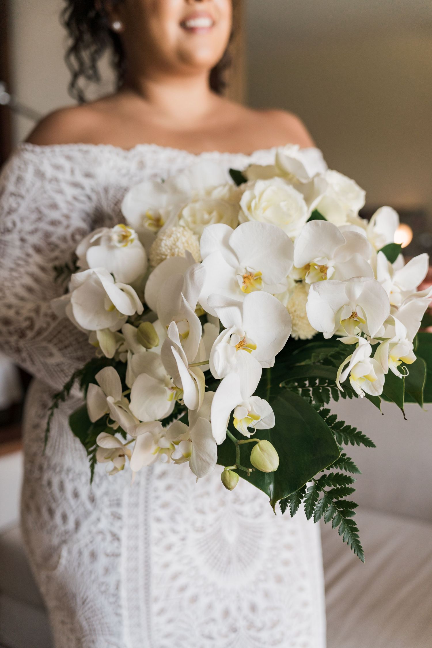 Xcaret Arte Wedding - Bride in white lace off-shoulder gown holding a lush bouquet of white orchids and roses with green ferns.