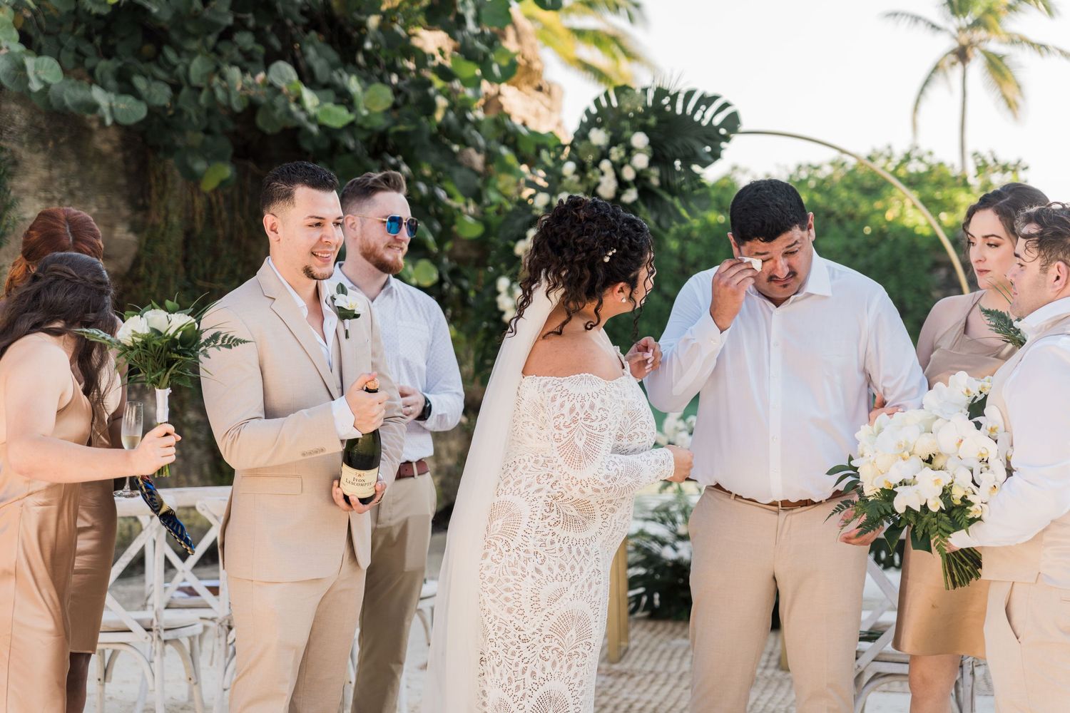 Xcaret Arte Wedding - Wedding party in tan suits and white dress laughing outdoors at a tropical beach ceremony with floral arch.