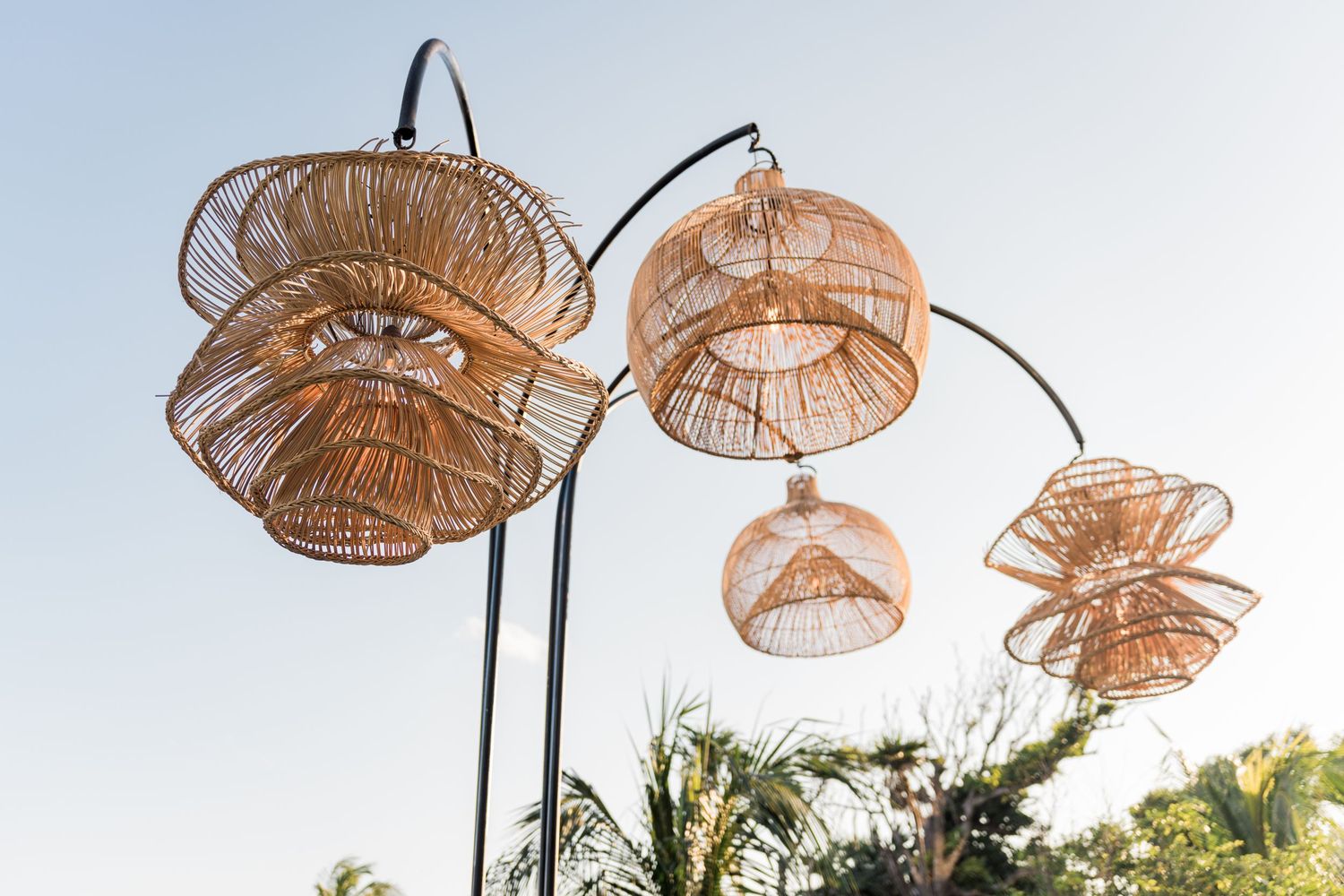 Xcaret Arte Wedding - Decorative wicker rattan pendant lamp shades hanging from curved black metal poles against a clear blue sky.