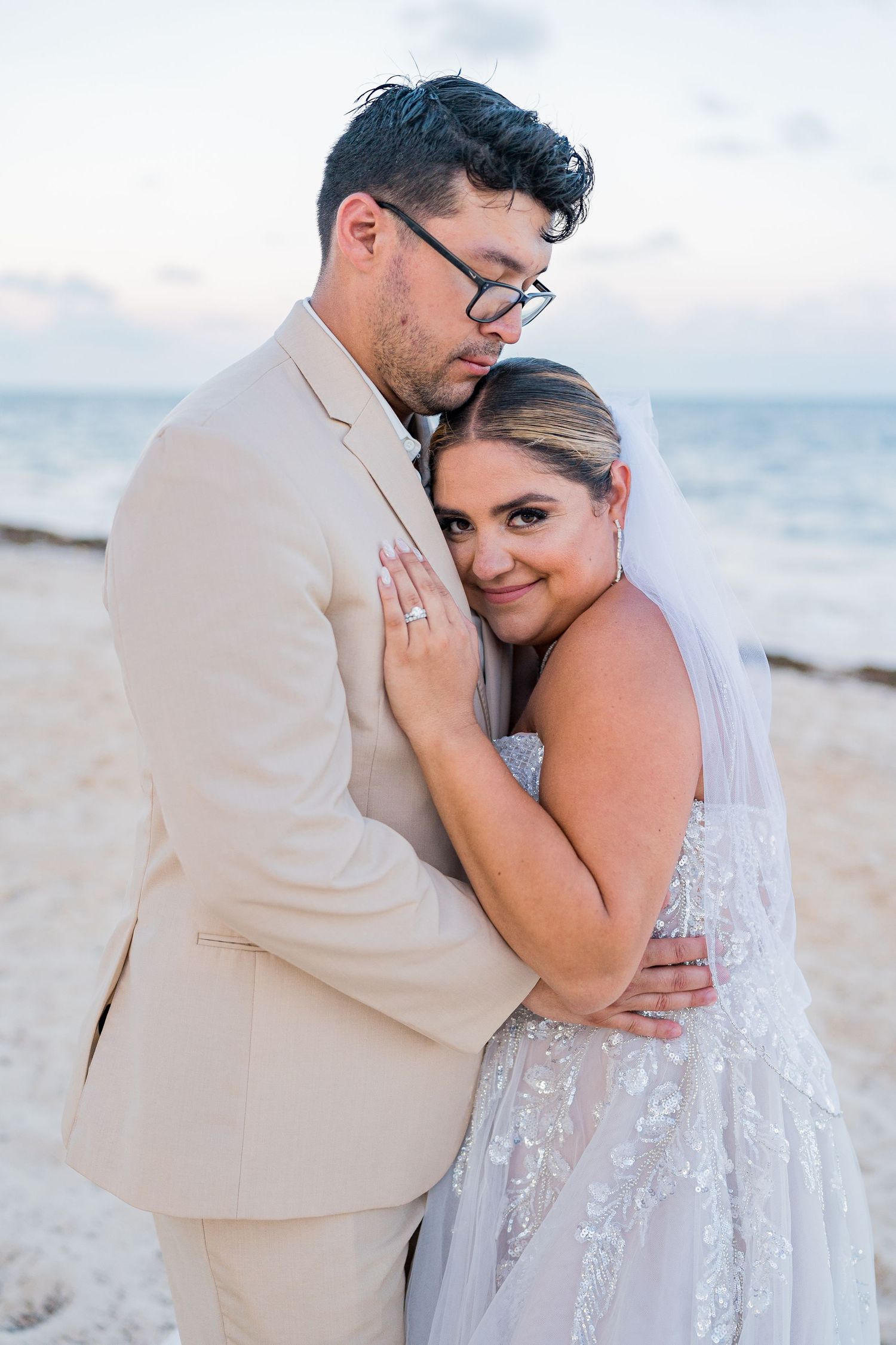 Bride leaning against groom in beige suit for intimate beach portrait at twilight.