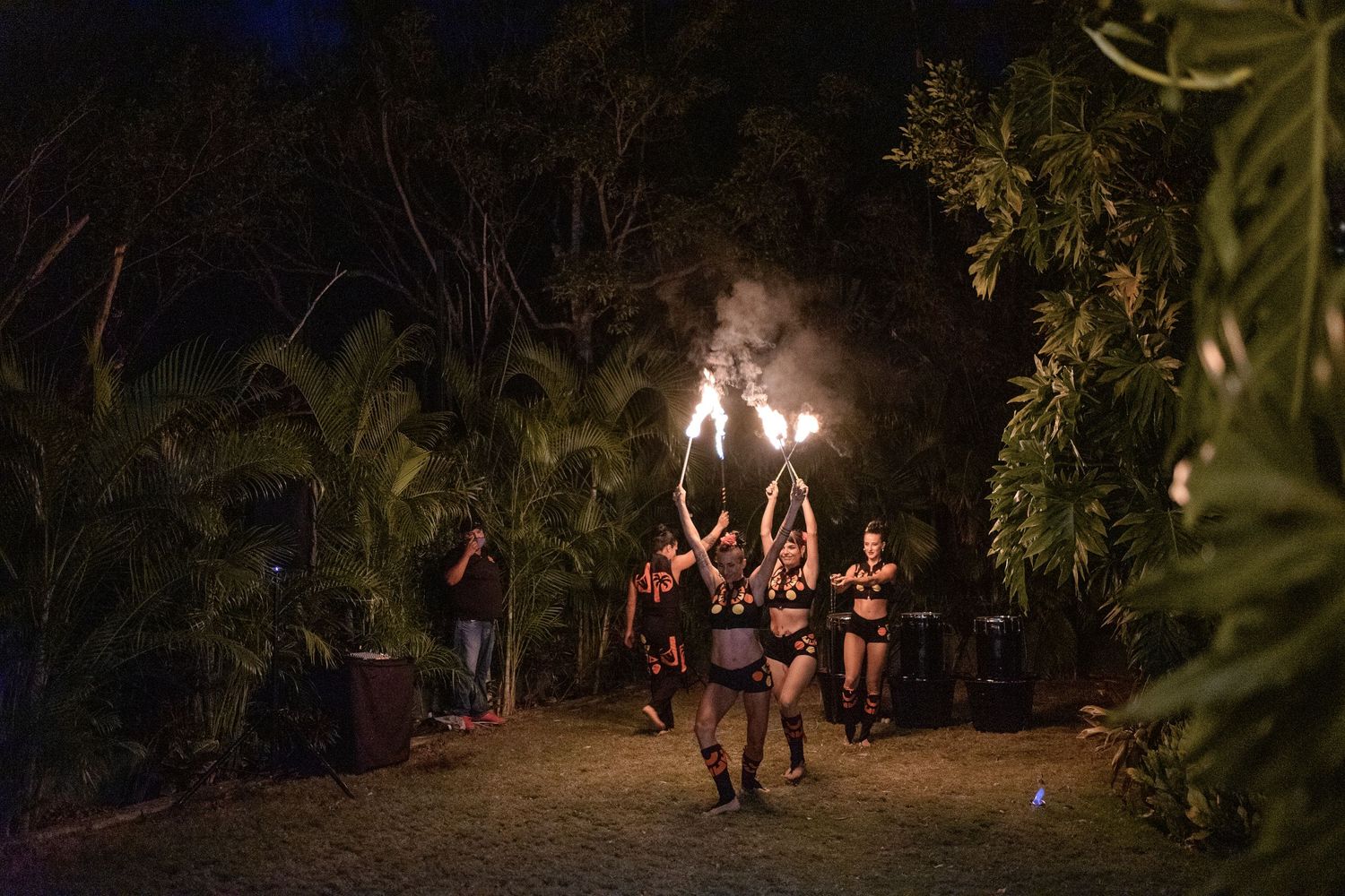 Group of fire dancers performing with flaming torches along a tropical garden pathway at night.