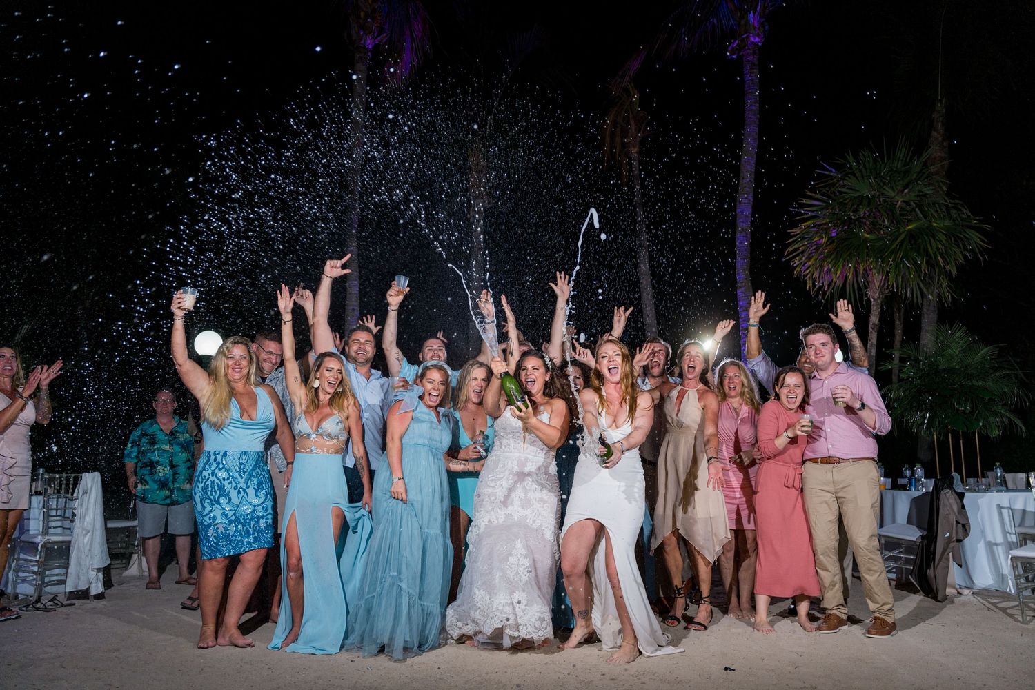 Wedding party celebrating with champagne spray on beach at night with palm trees.