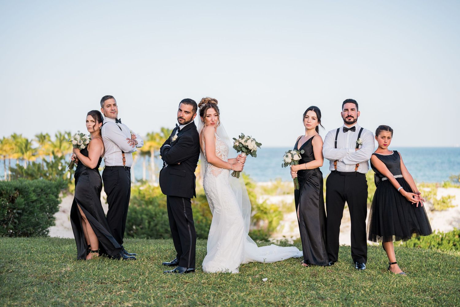 Elegant wedding party poses in pairs on seaside lawn, bridesmaids in black and groomsmen in suspenders.