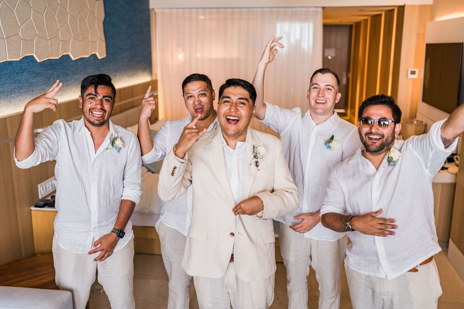 Groom and groomsmen in white suits celebrating together in hotel room before wedding.