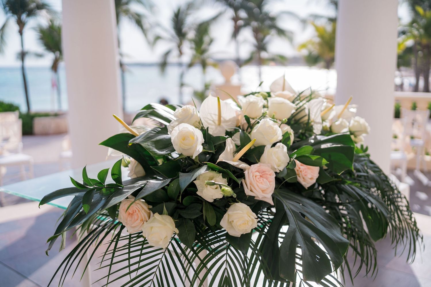 Elegant floral arrangement of white and blush roses with tropical greenery at a beachfront wedding ceremony.
