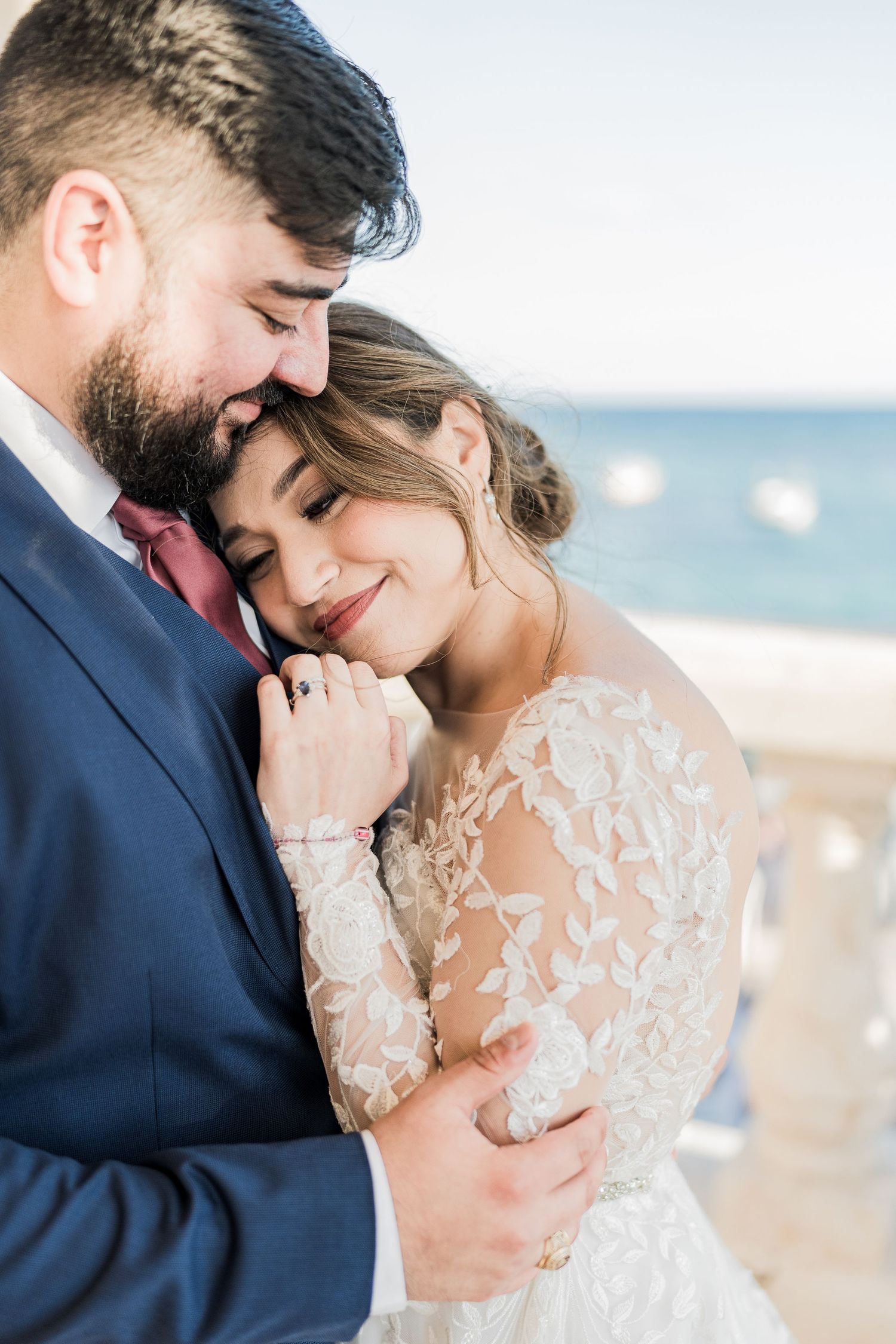 Bride in lace long-sleeve gown smiling while embracing groom in navy suit with ocean backdrop.