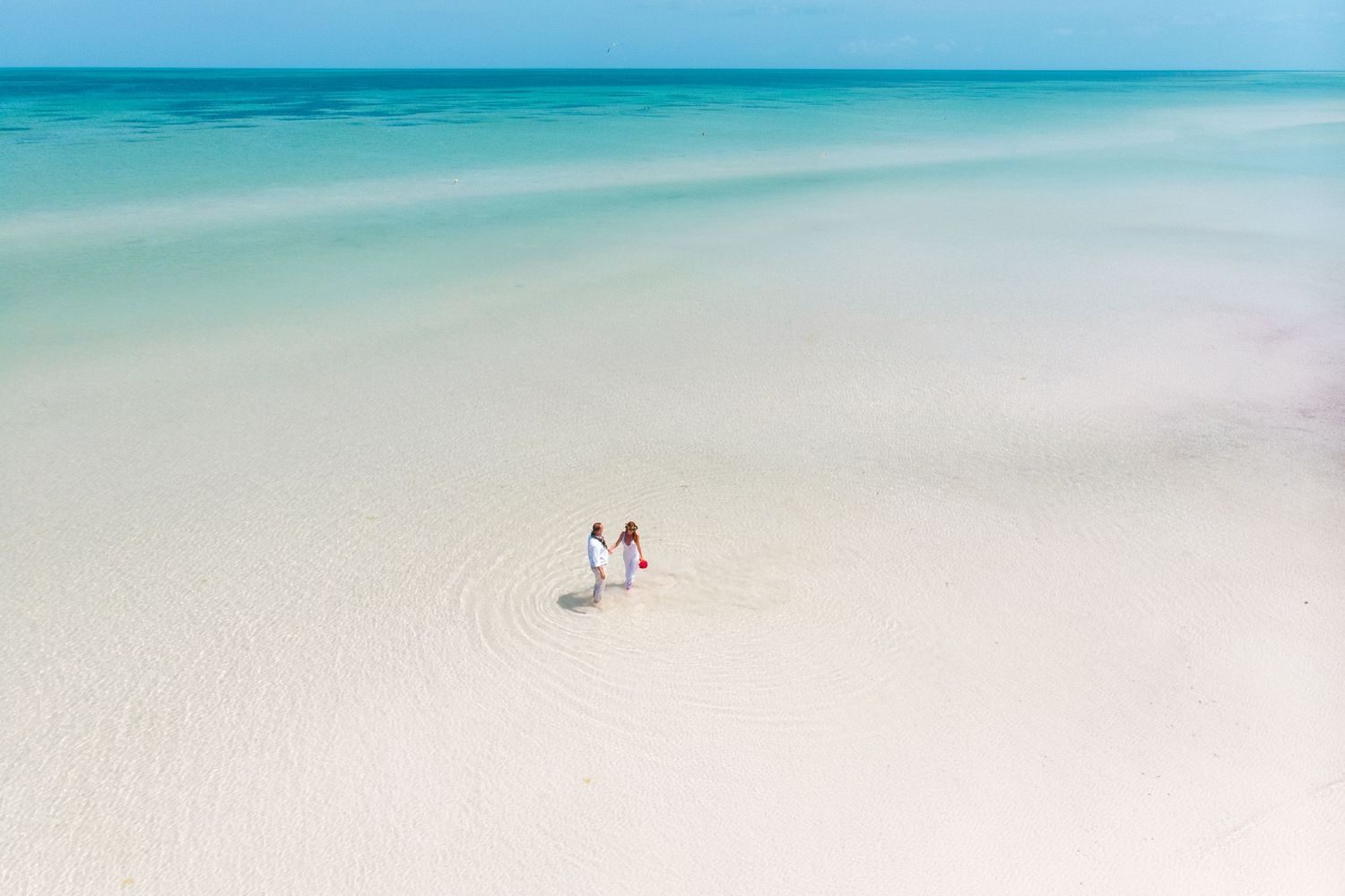 Aerial view of bride and groom walking on a pristine white sand beach with turquoise water.