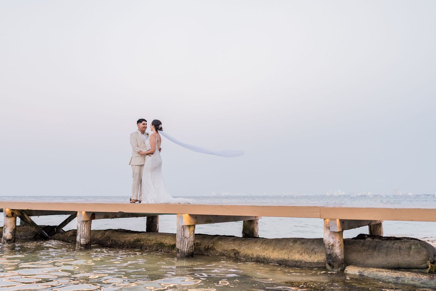 Bride and groom stand romantically on a wooden pier over calm ocean waters at their beach wedding.