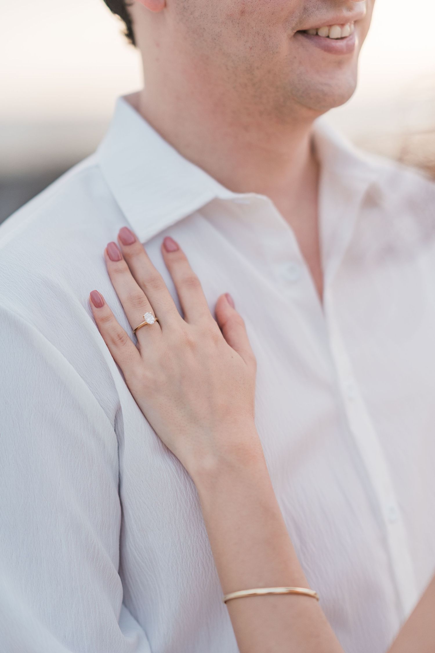 Close-up of a woman's hand with an engagement ring and gold bracelet resting on a man's white shirt.