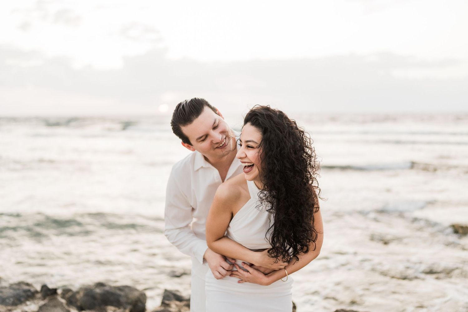 Couple laughing and embracing on a rocky beach at sunset, man in pink shirt hugging woman in white dress.