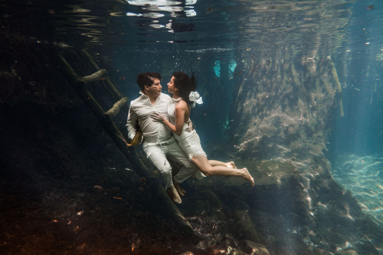 Couple in white clothing poses romantically underwater near a wooden ladder in a clear blue cenote.