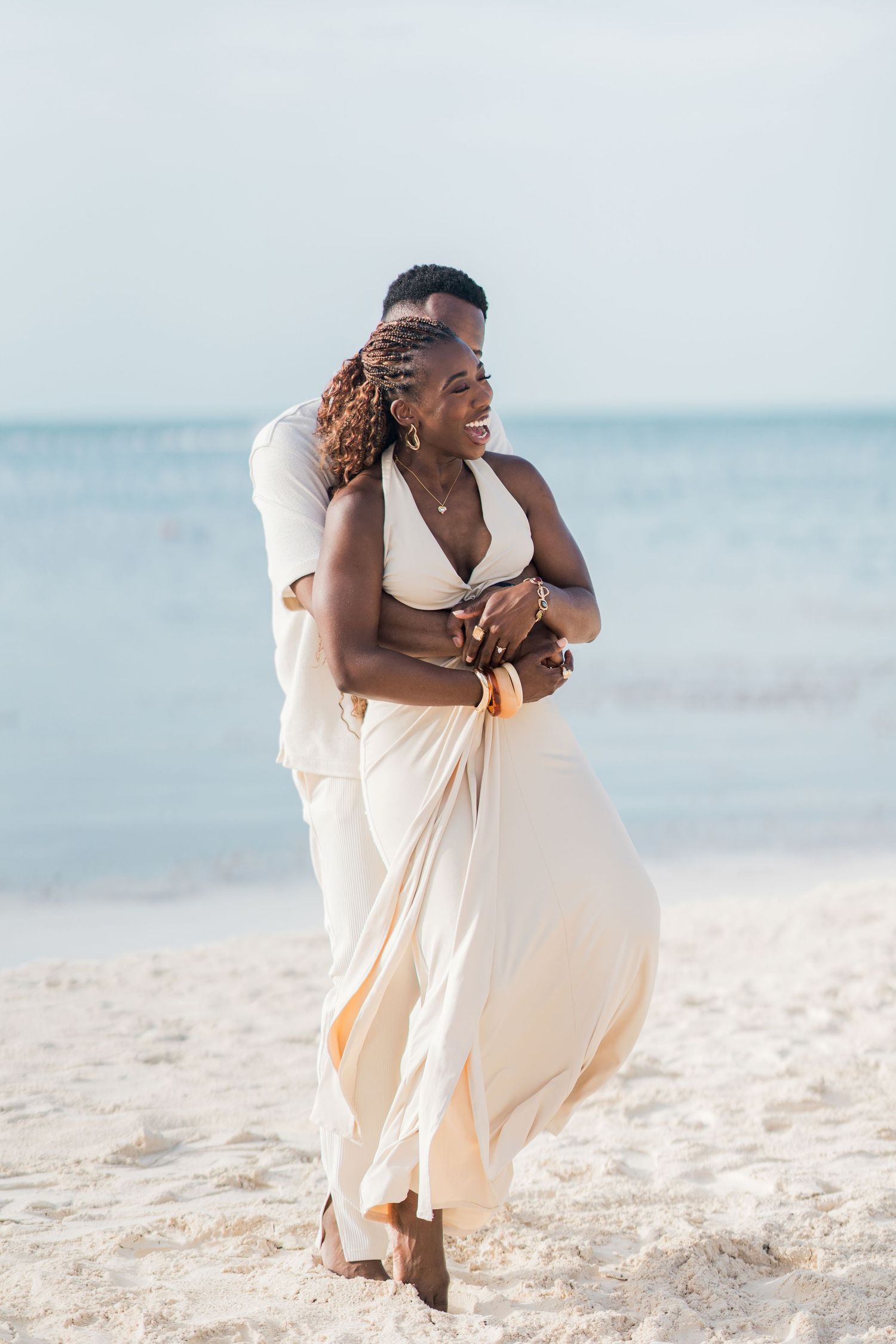 Joyful Black couple embracing on a sunny beach, woman in flowing white halter dress.
