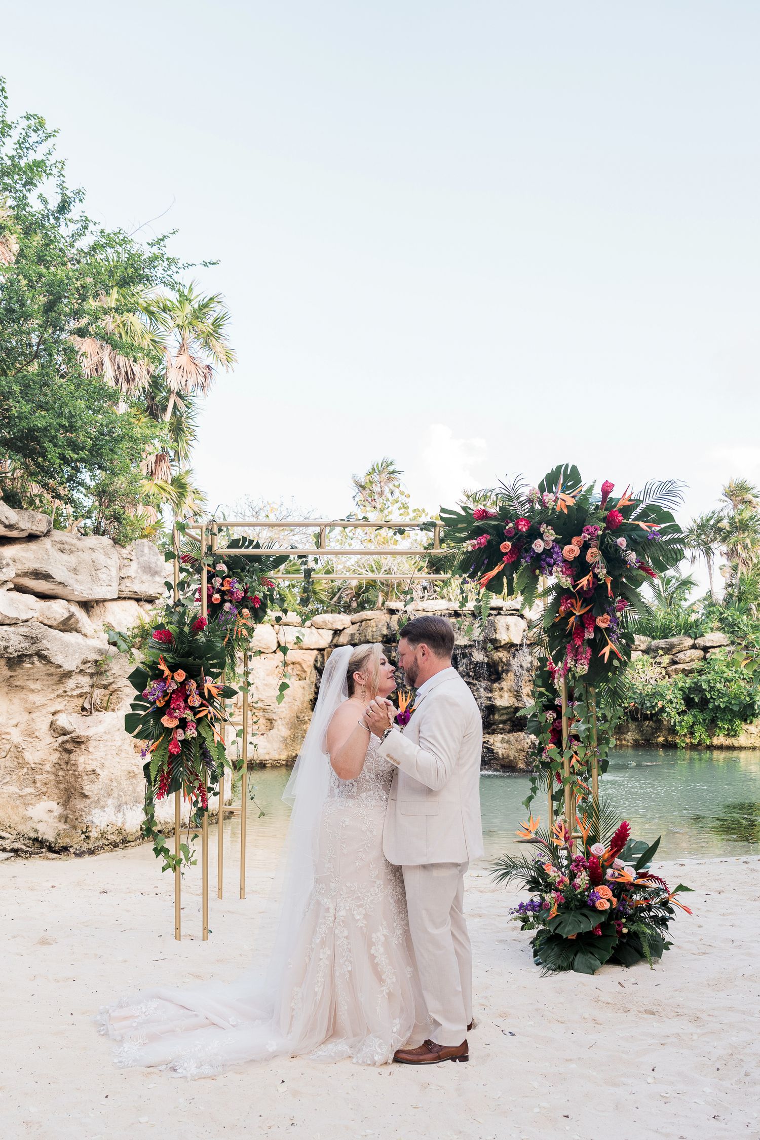 Bride and groom share first kiss under tropical floral arch on sandy beach at destination wedding ceremony.