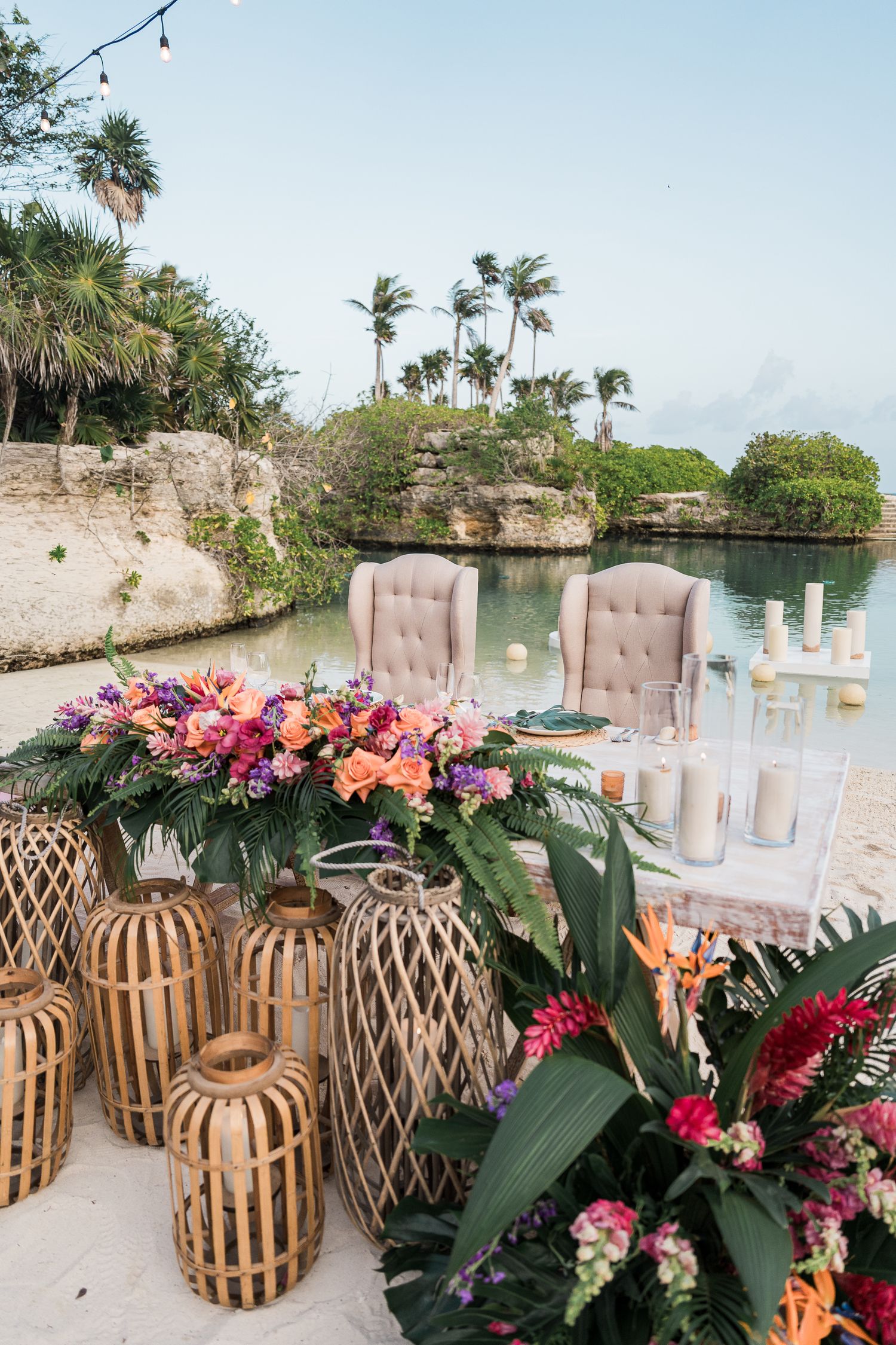 Hotel Xcaret México Tropical wedding reception table with colorful floral arrangements, and tufted chairs beside a serene lagoon.